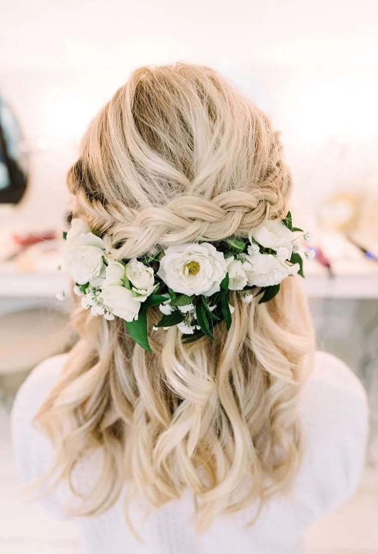 Back view of a woman with long, wavy blonde hair wearing a floral crown made of white flowers and green leaves, with a braided section in her hair.