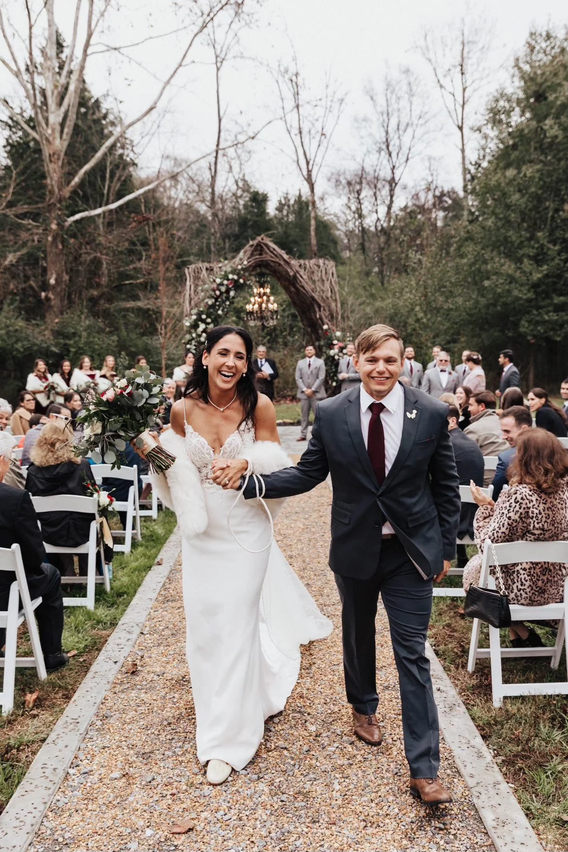 A bride and groom walking down the aisle after their wedding ceremony outdoors with guests seated on either side. The bride is holding a bouquet and smiling, wearing a white dress with a fur stole, while the groom is smiling, dressed in a dark suit a