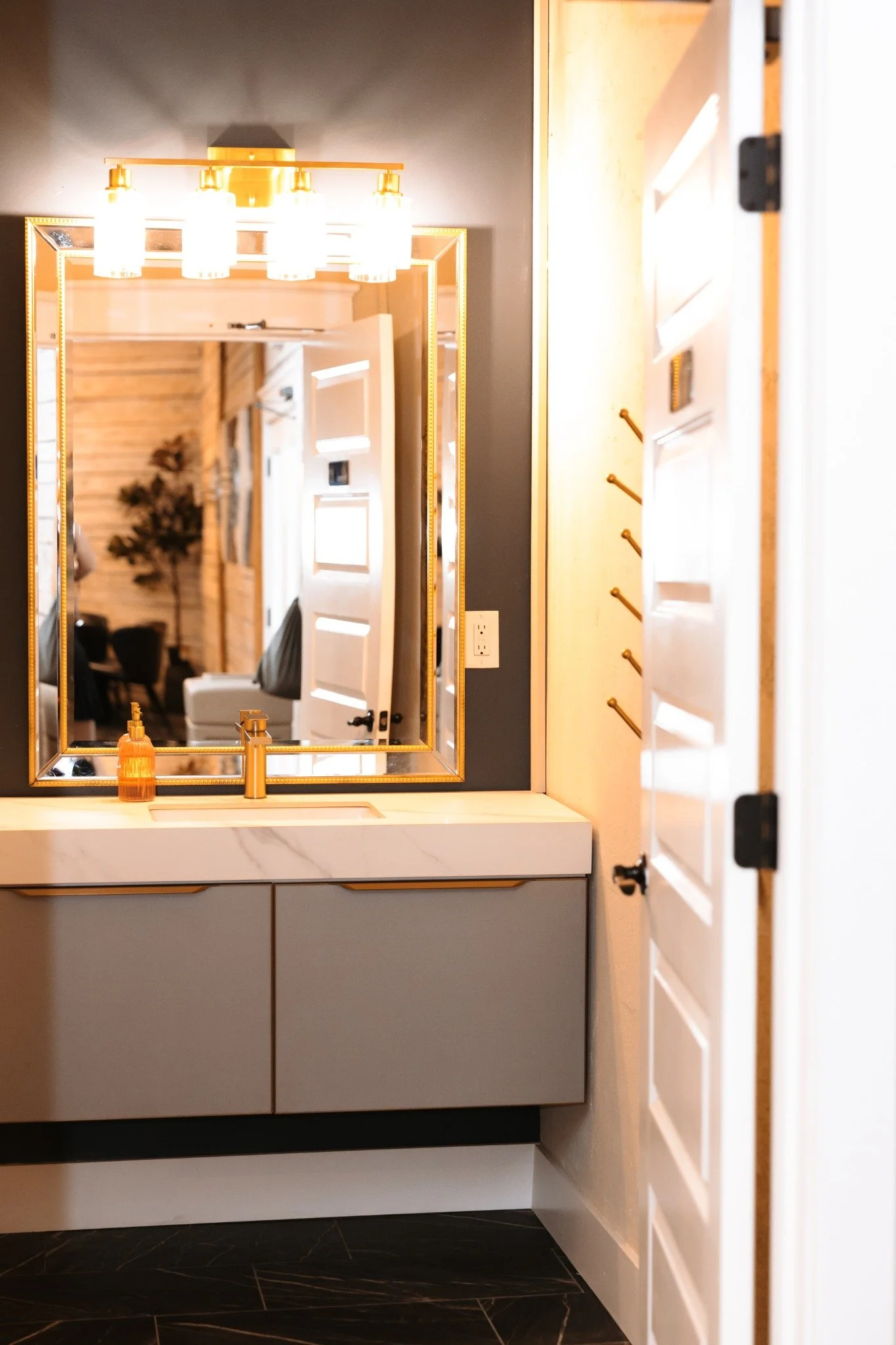 A bathroom mirror reflecting a modern vanity with gold fixtures and a white marble countertop, with a dark wall and wooden door.