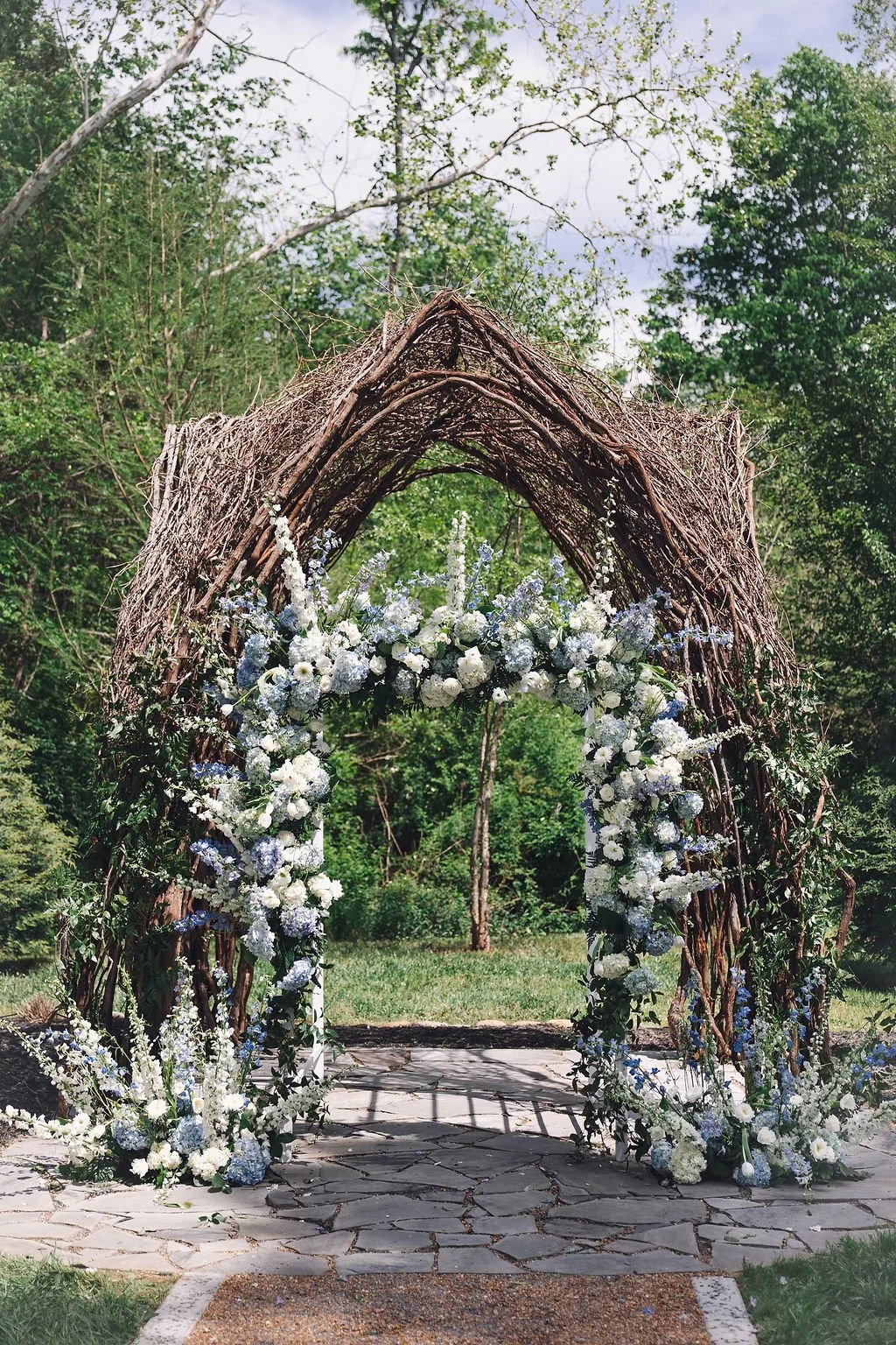Vine ceremony arbor decorated with cascading blue and white floral arrangements featuring hydrangeas, delphinium, roses, snapdragons, and lush greenery at Ramble Creek, styled for an outdoor garden wedding ceremony.