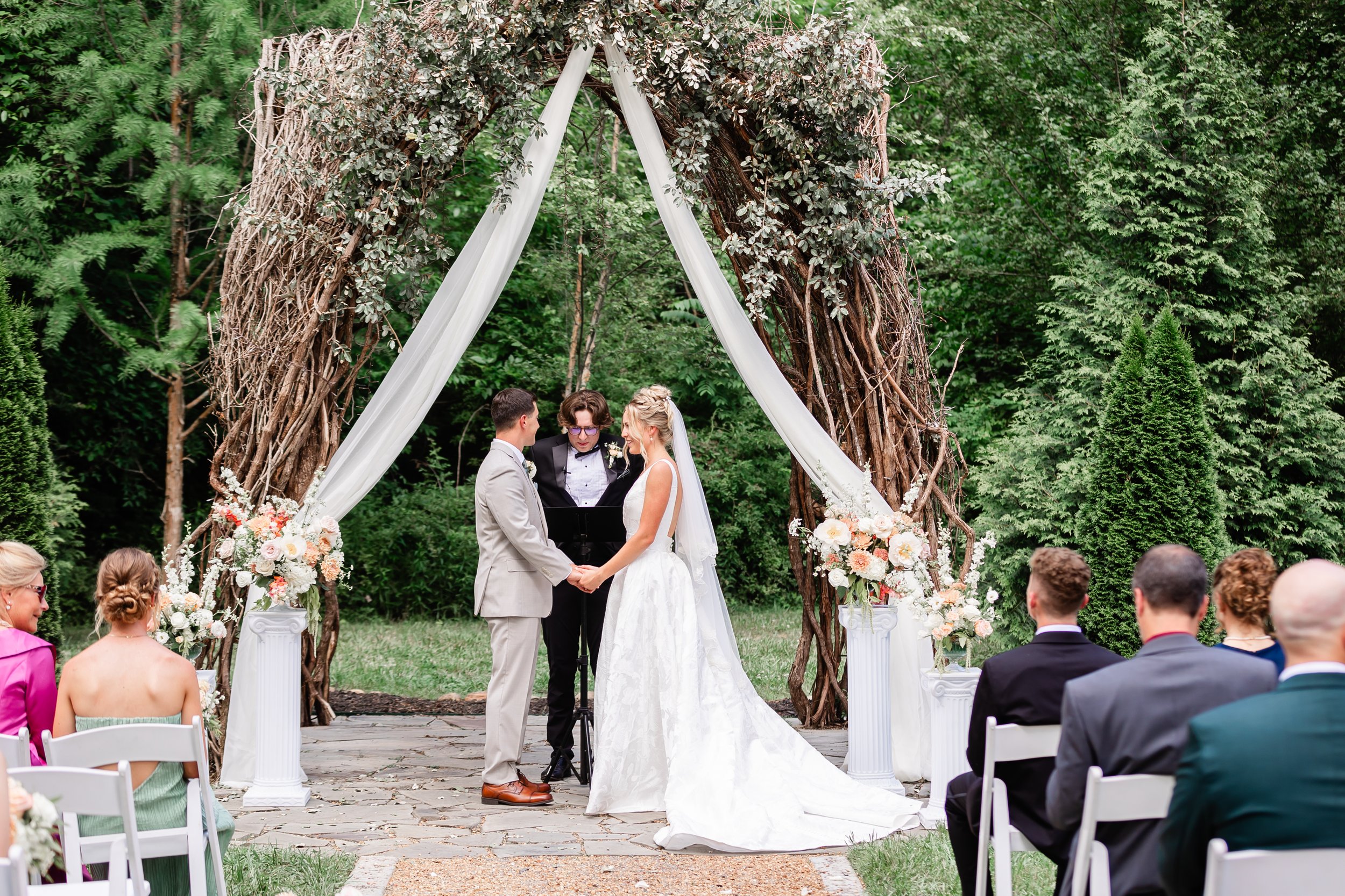 A Tennessee wedding ceremony outdoors with a couple exchanging vows under a wooden arch decorated with white fabric and flowers, surrounded by greenery and guests seated in chairs.
