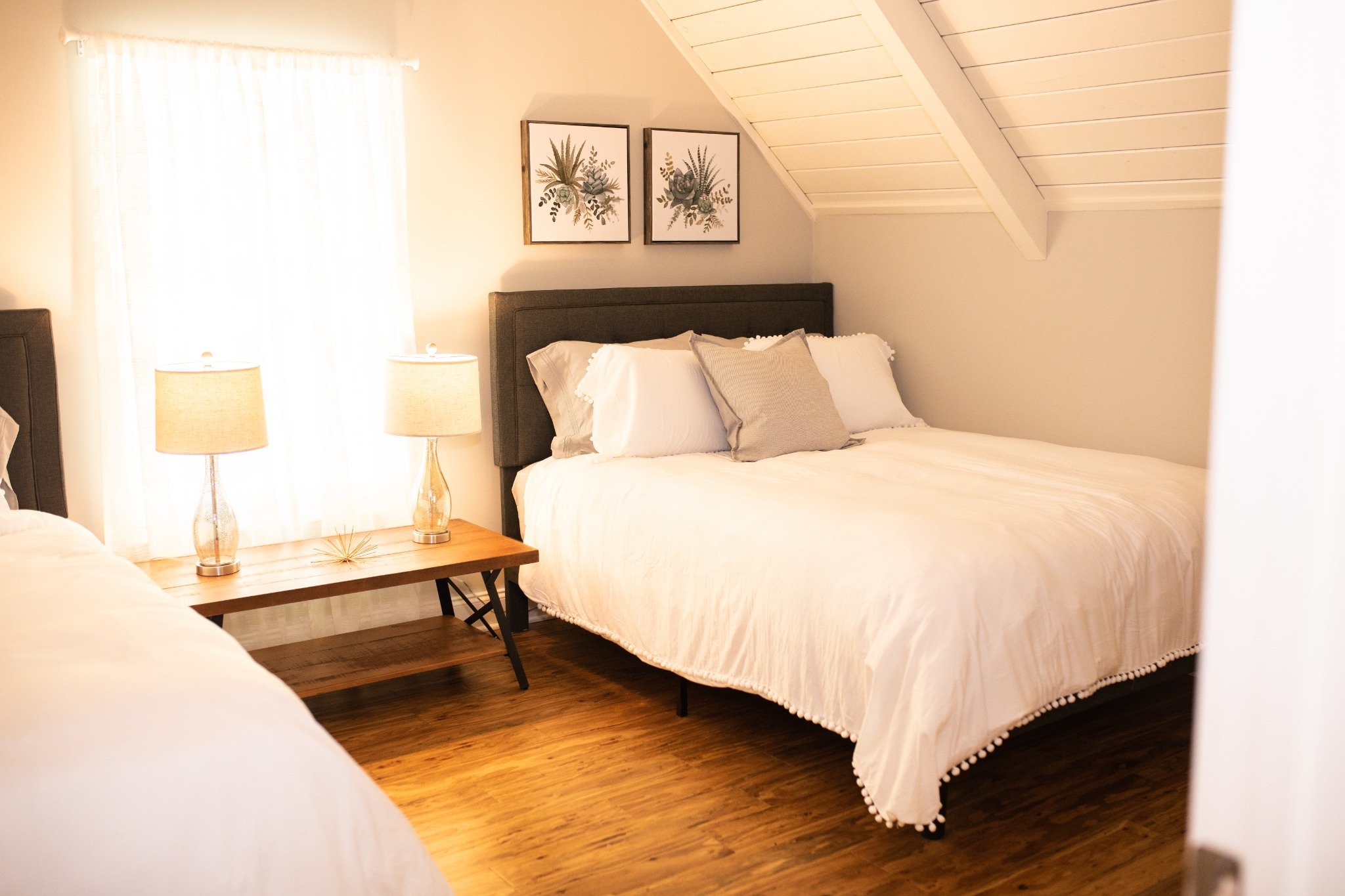 Upstairs bedroom at the Manor onsite accommodation at Ramble Creek, featuring white bedding, neutral accent pillows, bedside lamps, warm wood floors, and a cozy sloped ceiling.