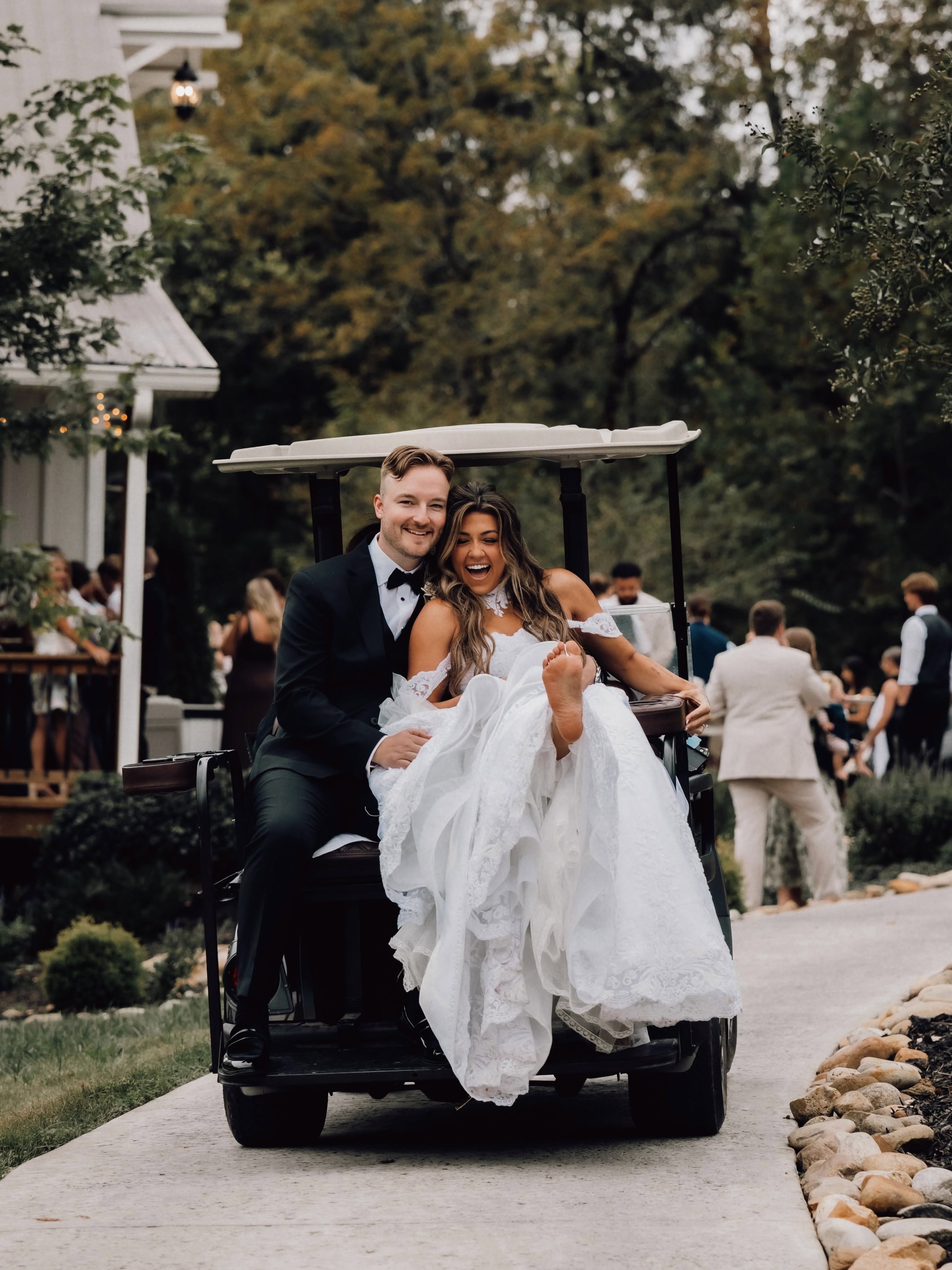 Kara Lewis and West Webb smile together on the back of a golf cart outside Ramble Creek’s Grand Hall, with Kara playfully lifting a bare foot beneath her wedding dress.