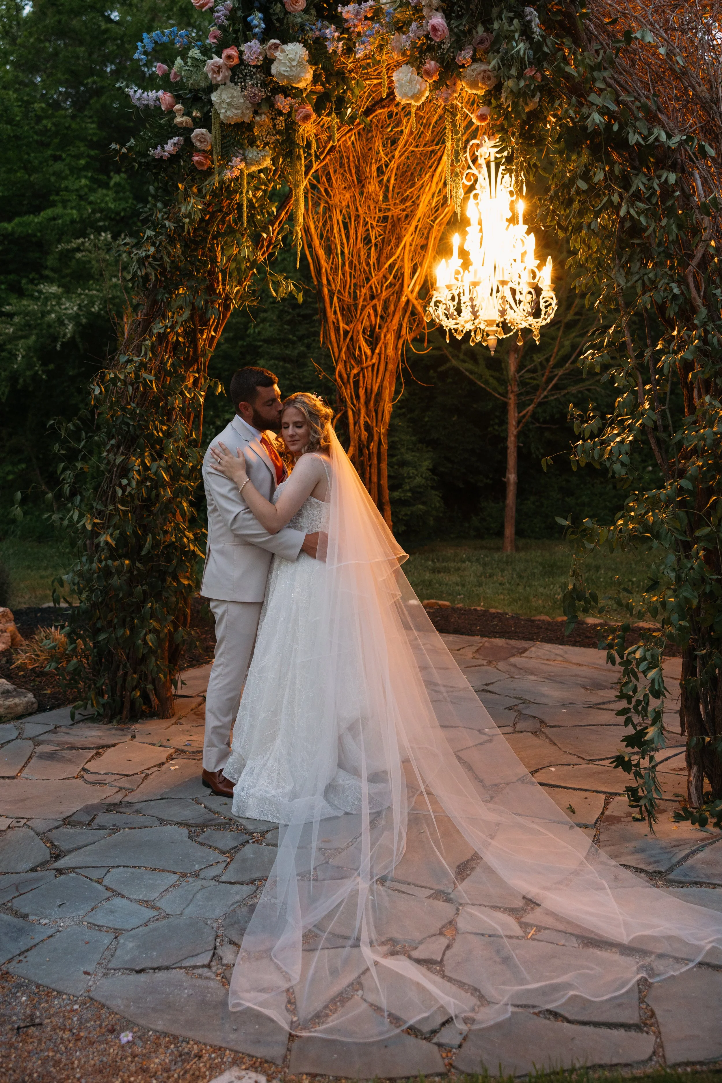 A bride and groom embracing under a decorative arch adorned with flowers and lit by a chandelier, outdoors during dusk on a stone patio.