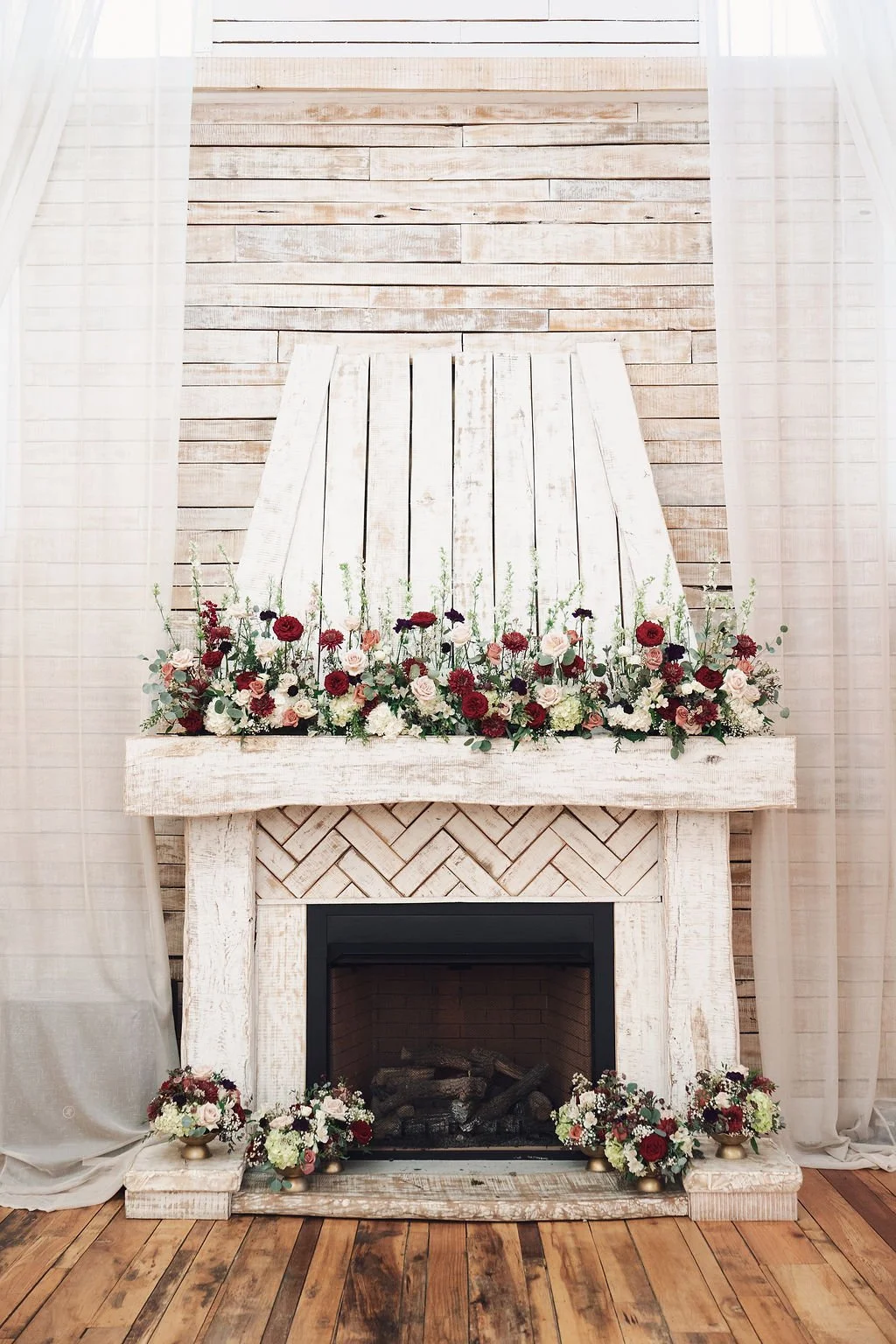 Styled fireplace with a whitewashed wood mantel and a floral arrangement with red, white, and pink flowers, backed by a light wood-paneled wall and flanked by white curtains, ready for indoor wedding ceremony or reception.