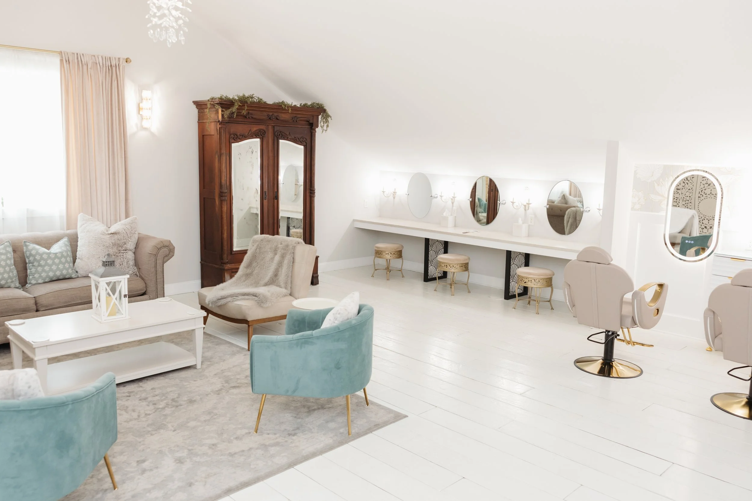 A bright, elegant room with white walls and floors, decorated with pastel-colored furniture. There are multiple round mirrors on the wall, a vintage wooden wardrobe, and a seating area with a beige sofa and turquoise armchairs. A white coffee table i