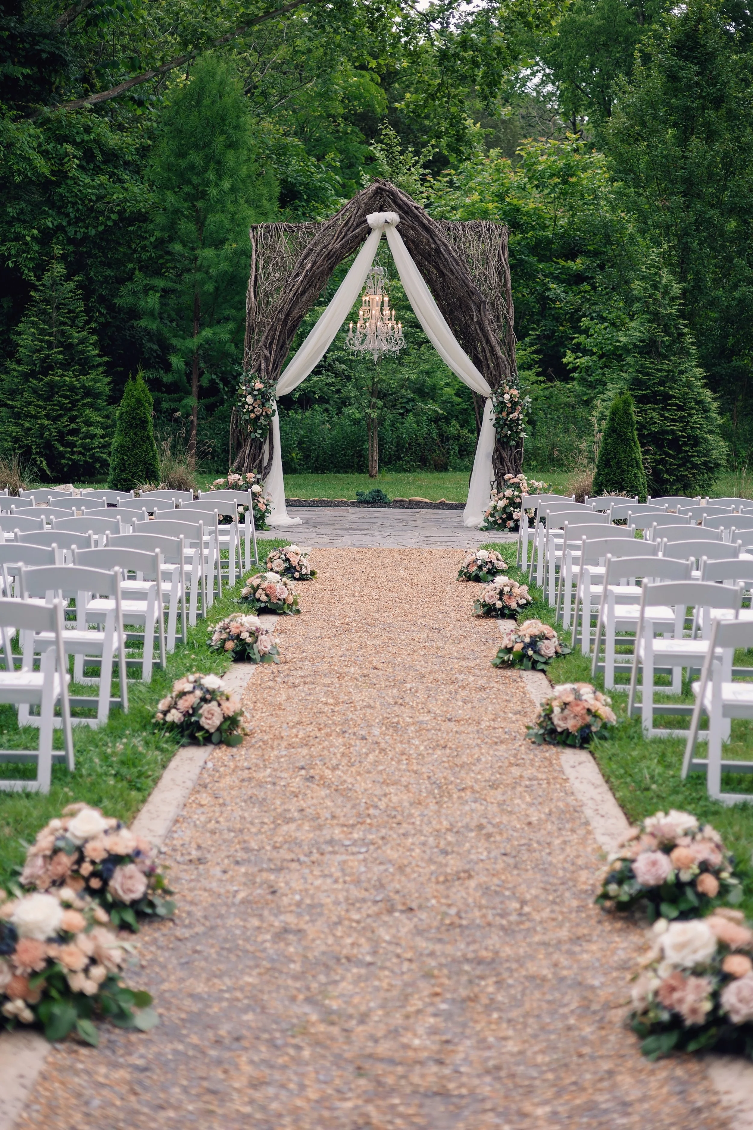 Wedding aisle lined with flower arrangements leading to a decorated archway with draped fabric and a chandelier, set outdoors in a lush green forest.