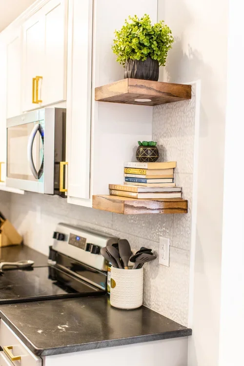 Kitchen corner with white cabinets, gold handles, microwave, stove, black countertop, wooden shelves holding a potted plant, books, and a container with kitchen utensils, and a white wall socket.