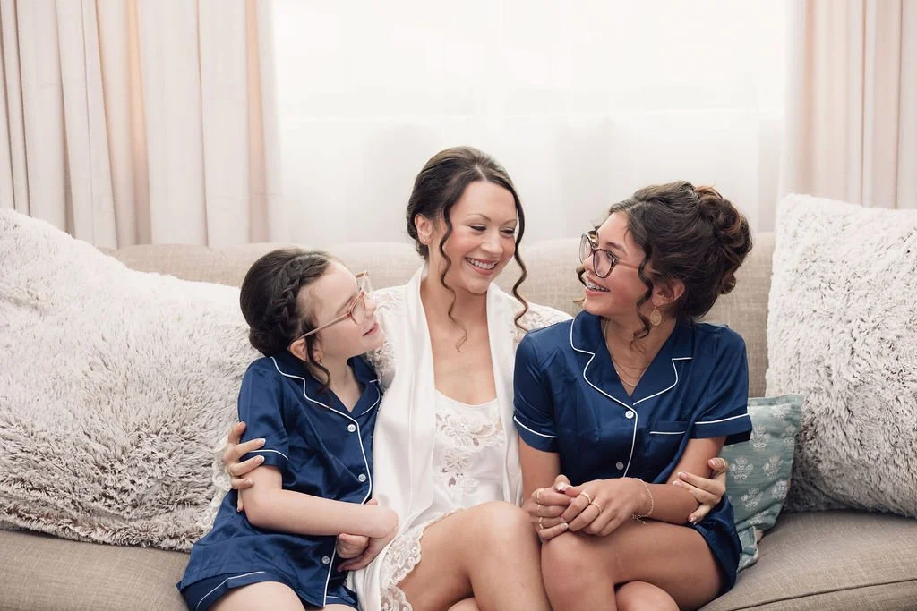 Bride smiling on a couch with two young flower girls in navy pajamas during wedding morning preparations.