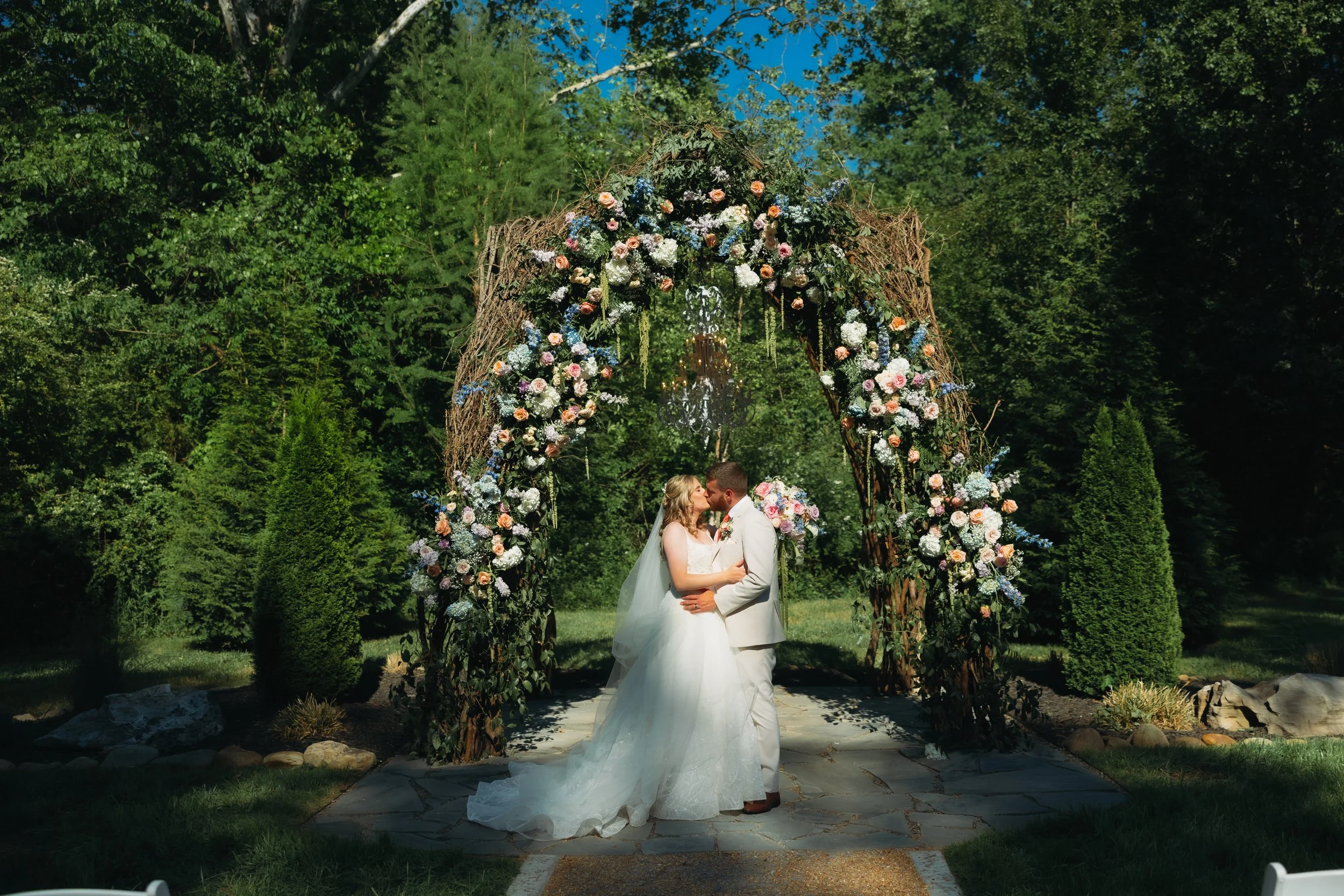 Bride and groom kissing beneath a lush outdoor wedding ceremony arbor covered in greenery, pastel flowers, and a chandelier in a garden setting