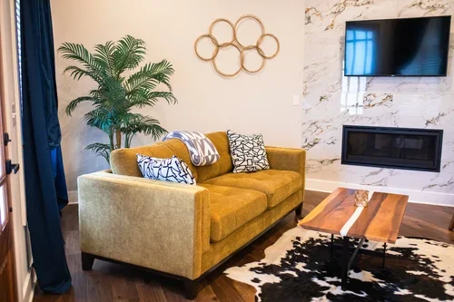 Living room with a yellow sofa, decorative pillows, a wooden coffee table, a large potted plant, a wall-mounted TV, and a marble accent wall. One of three accommodations at Ramble Creek Tennessee wedding venue with on-site lodging.