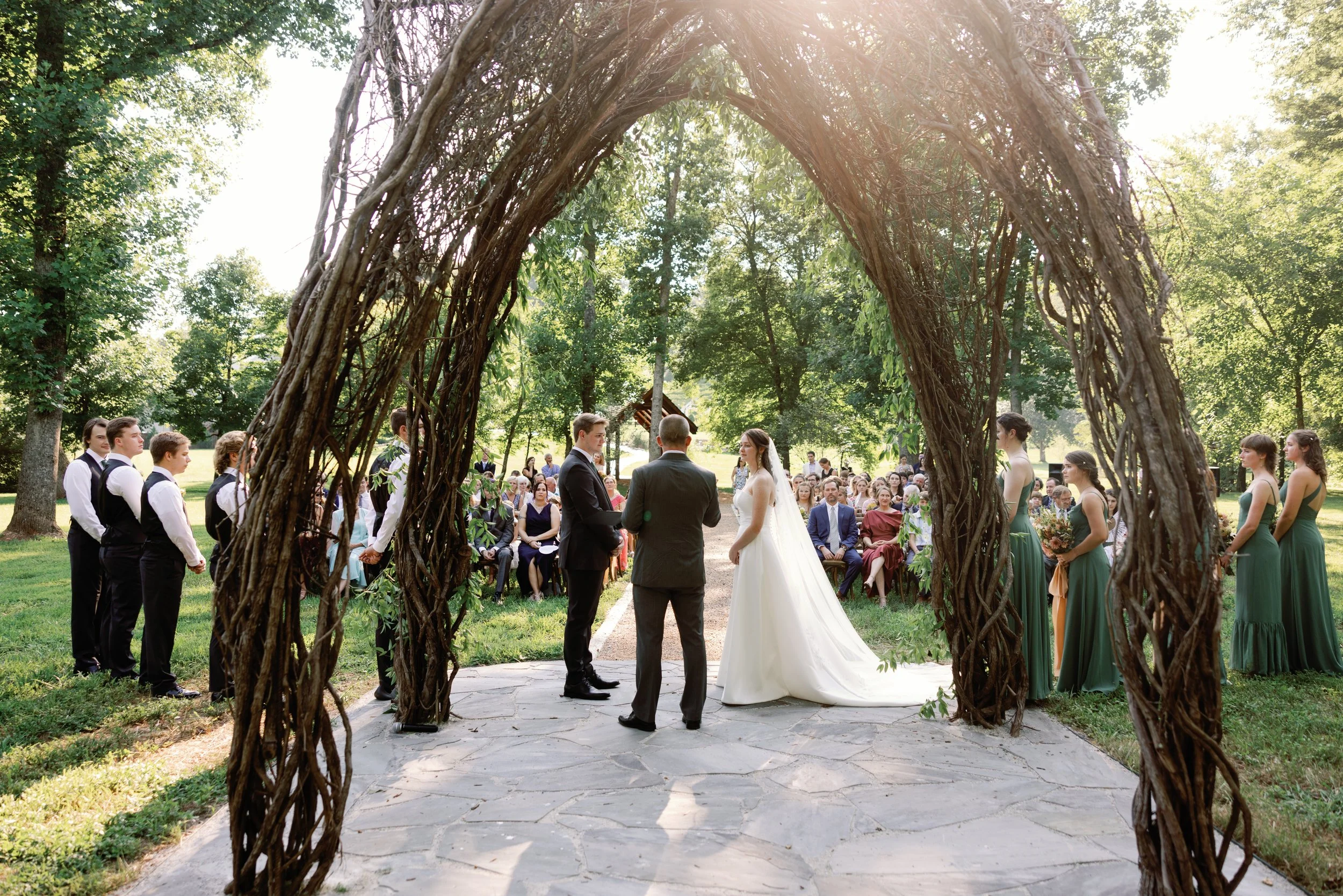 Outdoor wedding ceremony underneath a large woven arbor made of intertwined branches, with the bride and groom standing facing each other. Guests seated in the background surrounded by lush green trees, and sunlight filters through the leaves.
