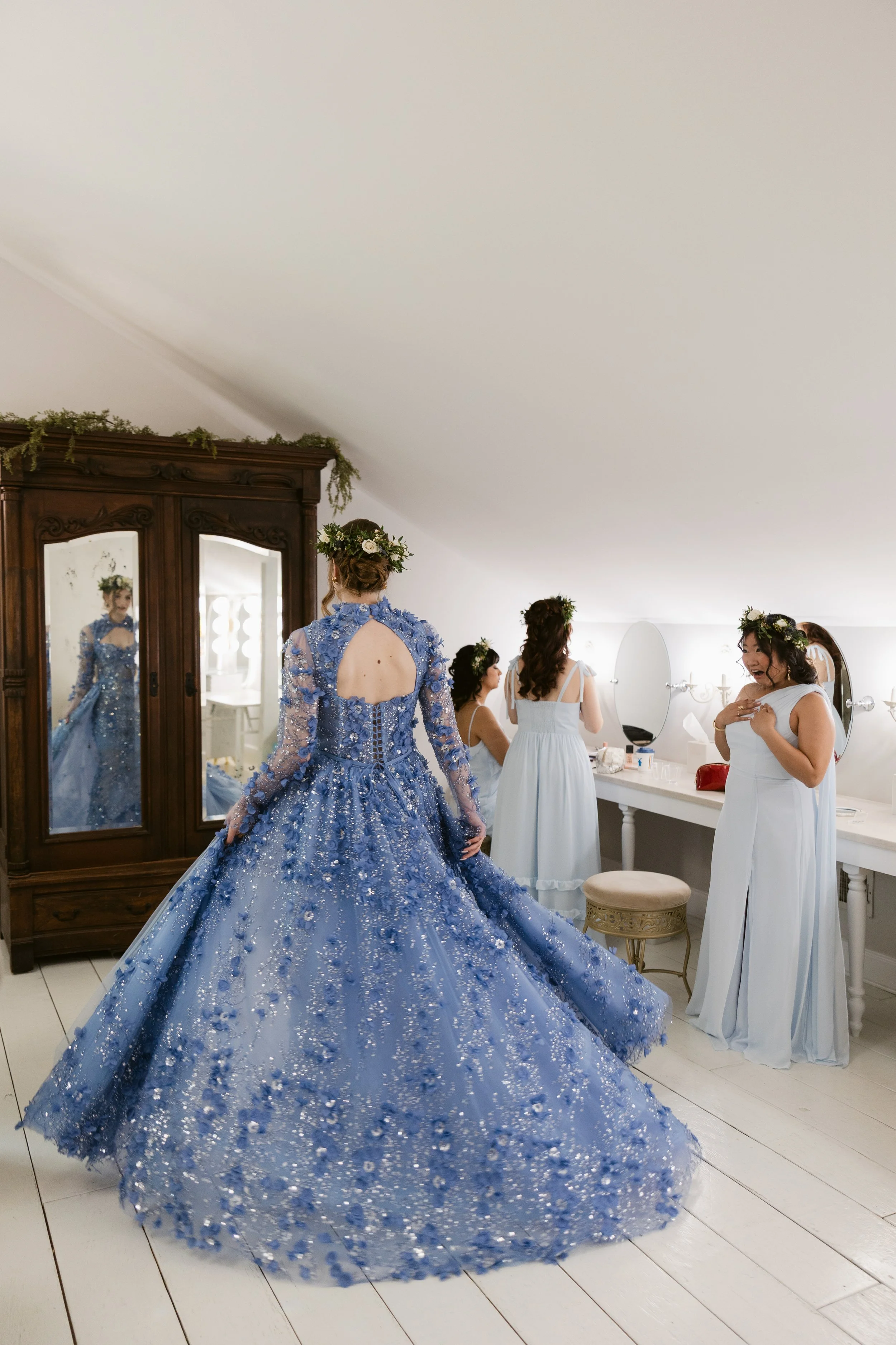 A bride in a large, non-traditional ornate blue gown with floral details and a floral crown on her head, standing in front of a mirror while bridesmaids in white dresses prepare in a bridal sui with white walls, a wooden wardrobe, and a vanity table.