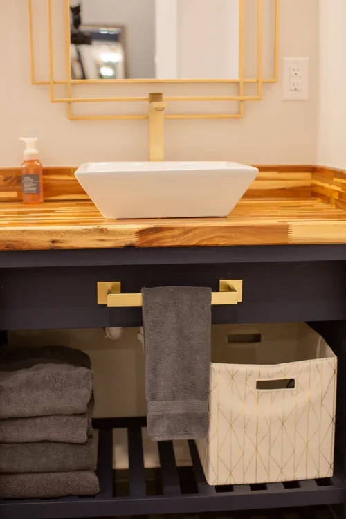 Bathroom vanity with a wooden countertop, white vessel sink, gold faucet, mirror with gold frame, soap dispenser, hand towel on gold bar, and storage baskets underneath. One of three accommodations at Ramble Creek Tennessee wedding venue with on-site