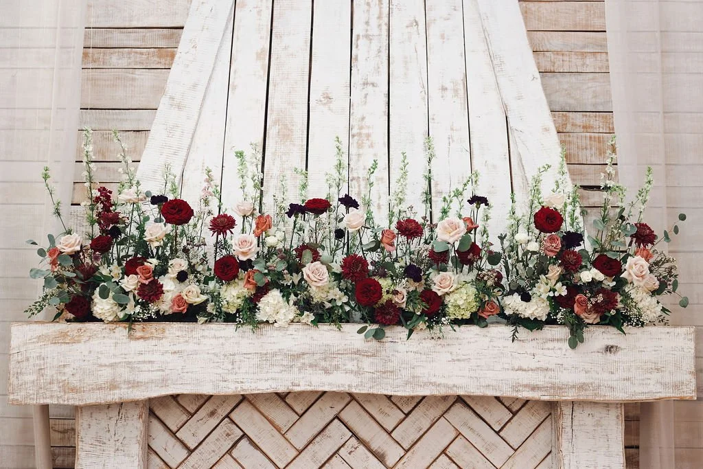 Fall and winter wedding mantel floral arrangement with red, blush, ivory, and burgundy flowers against a whitewashed wood backdrop
