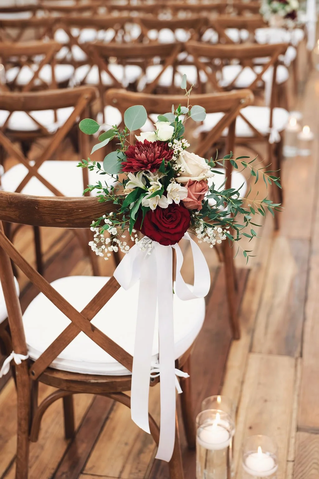 Fall wedding ceremony chair decorated with red, blush, and white flowers, greenery, and a white ribbon along the aisle
