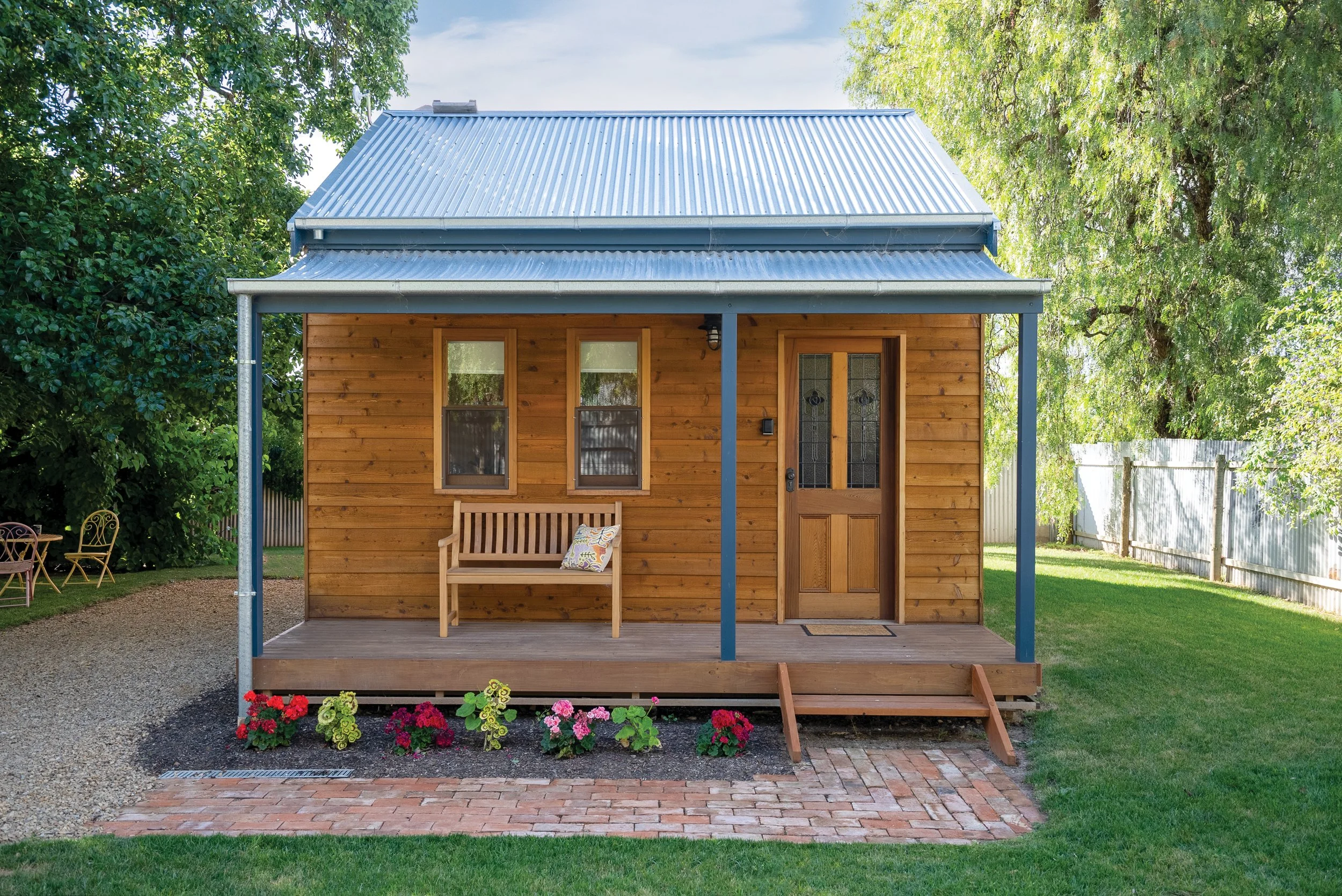 A wooden cabin at Tread Lightly Willunga with a metal roof, front porch, and two windows, surrounded by trees with colorful flowers.