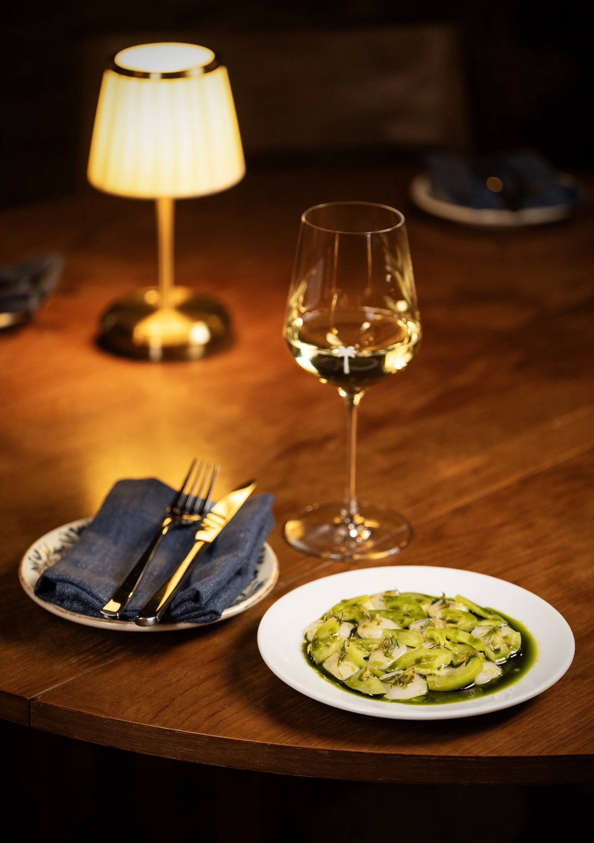 A glass of white wine, a lit table lamp, a plate of scallop crudo, and silverware on a dark wooden table in the Cellar at Tread Lightly.