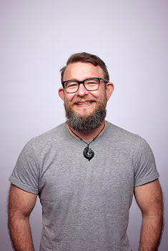 A man with glasses and a beard smiling, wearing a gray t-shirt and a necklace, standing against a light-colored background.
