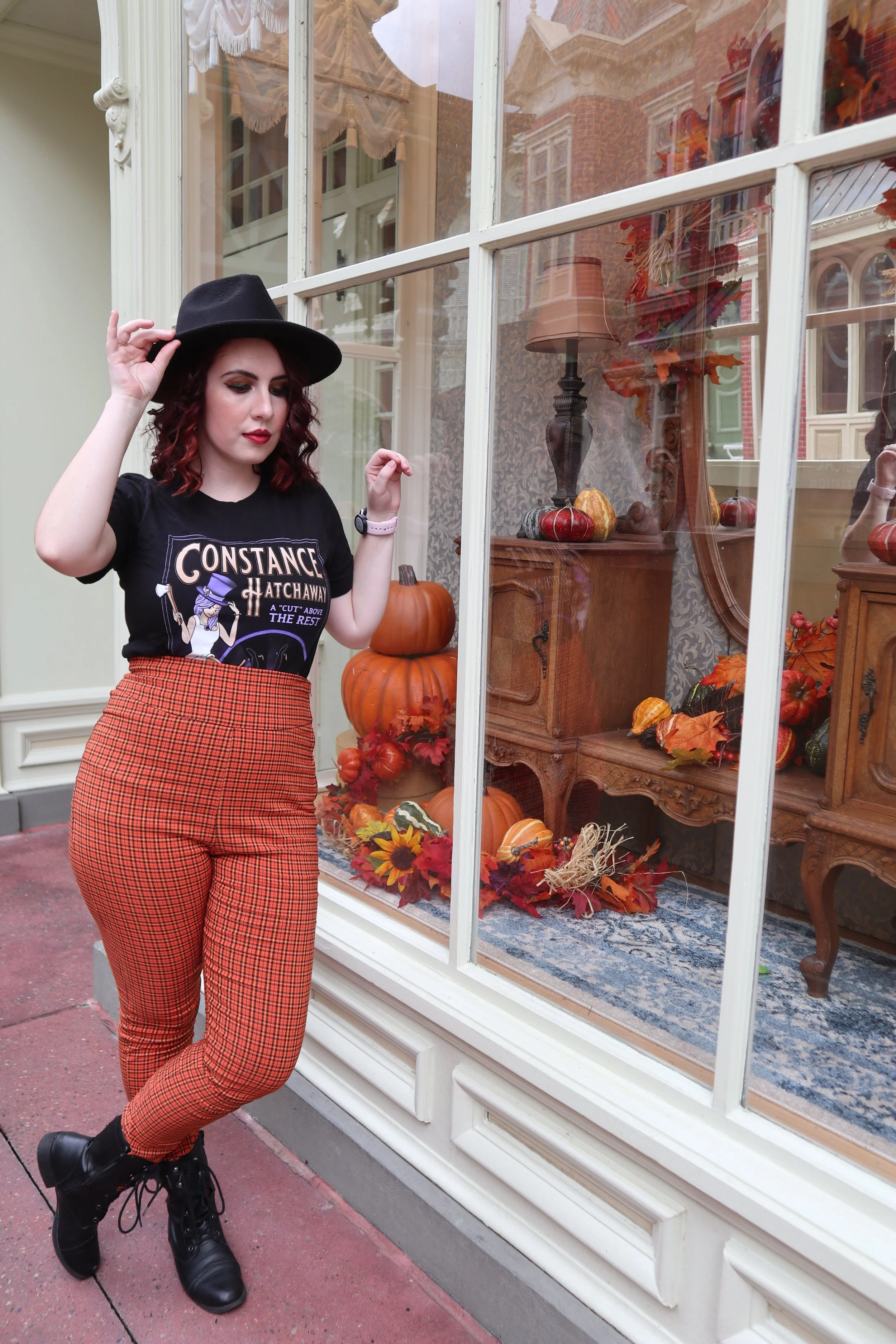 Woman with wavy dark red hair, wearing a black hat, black graphic t-shirt, orange plaid pants, and black boots, standing outside a display window decorated for fall with pumpkins, gourds, leaves, and an indoor lamp with a beige shade.