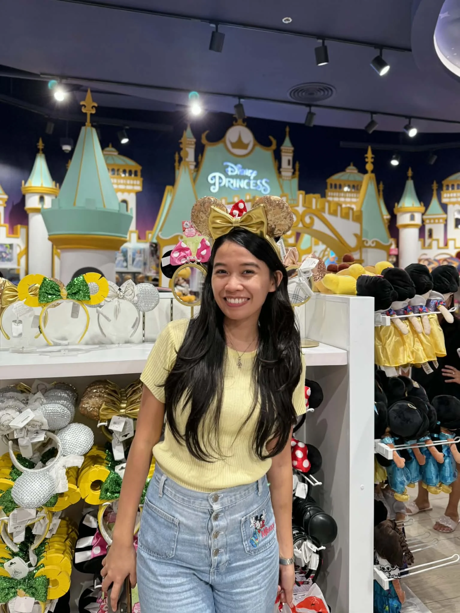 A woman wearing a yellow top and denim shorts stands in a Disney-themed merchandise store, with Disney Princess castle background and various headbands and plush toys around her.