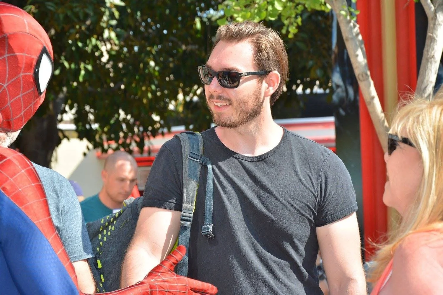 A man wearing sunglasses and a black t-shirt talking to two women, one of whom is wearing sunglasses, outdoors with trees and a red structure in the background.