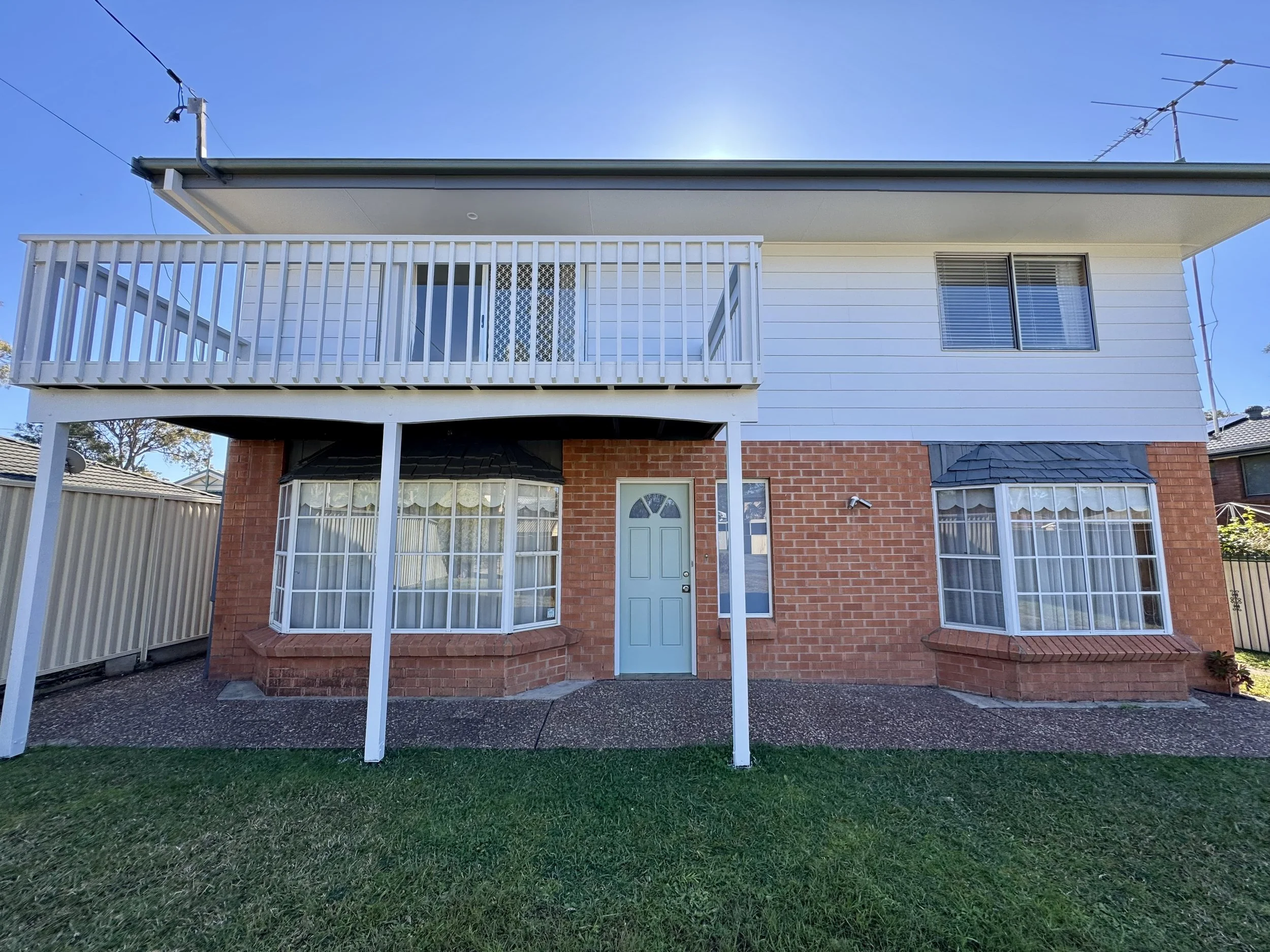 A two-story house with red brick on the lower level and white siding on the upper level. It features a small balcony with white railing and a light blue door. The backyard has a green lawn and a gravel path, with a neighboring house visible to the right and trees in the background.