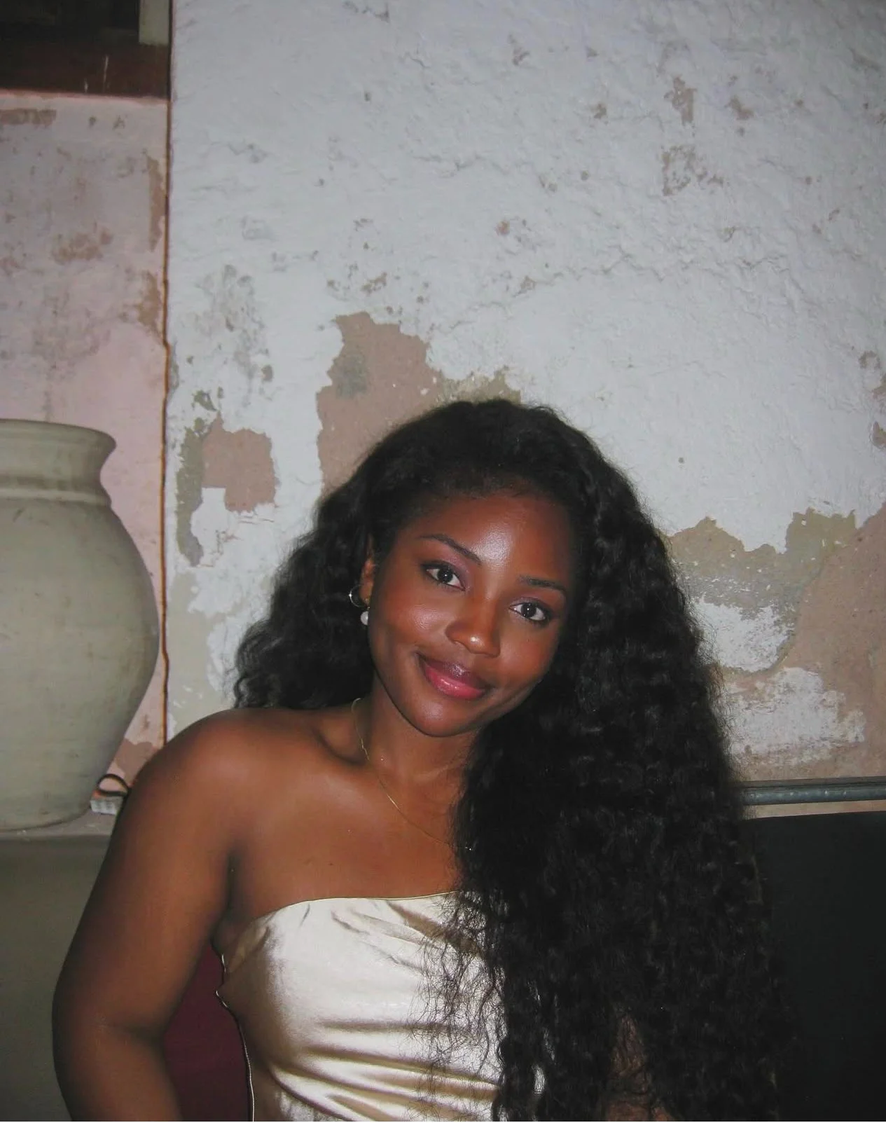 A young woman with long curly black hair, wearing a strapless white top, smiling at the camera against a weathered wall with peeling paint and a large ceramic pot to her left.