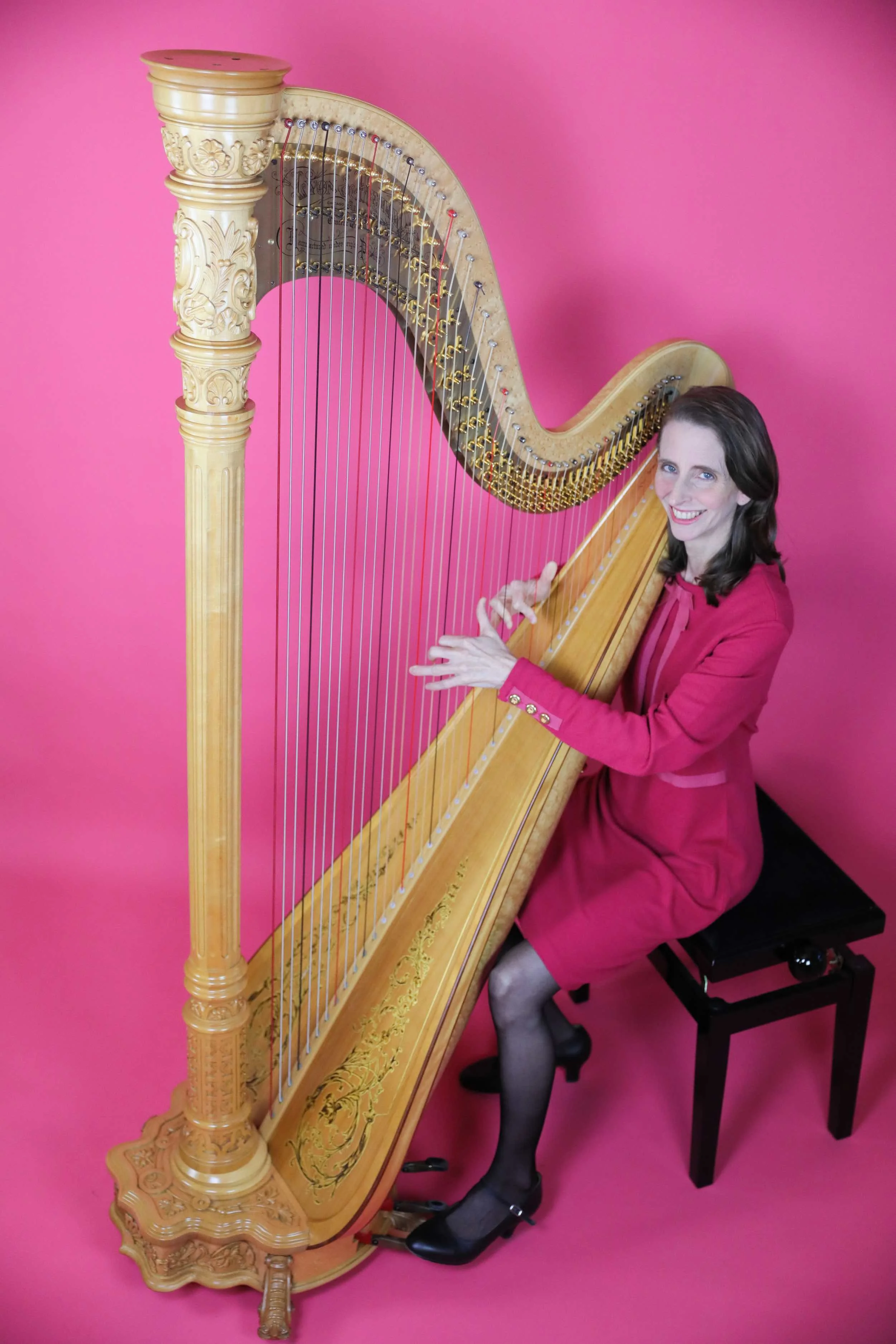 A woman in a pink blazer and skirt is sitting on a small black stool playing a large concert harp with intricate carvings. The background is plain pink.