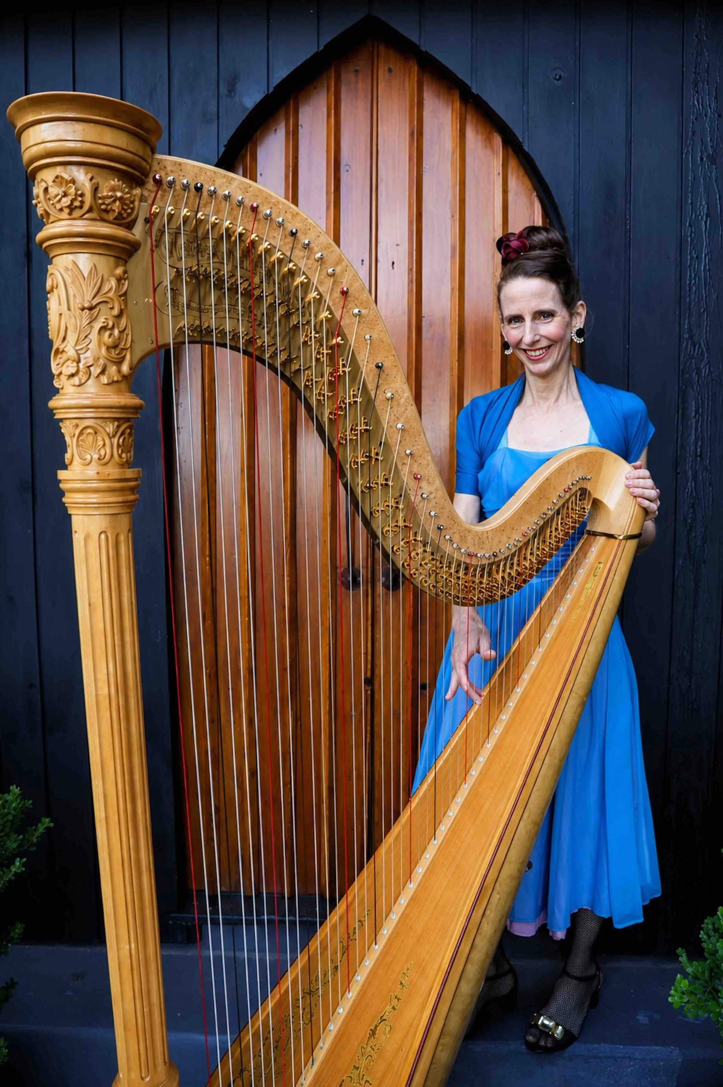 A smiling woman dressed in vintage style clothing, with a blue dress, black shoes, and black stockings, standing beside a large concert harp with ornate carvings, against wooden church doors and a dark blue wall.