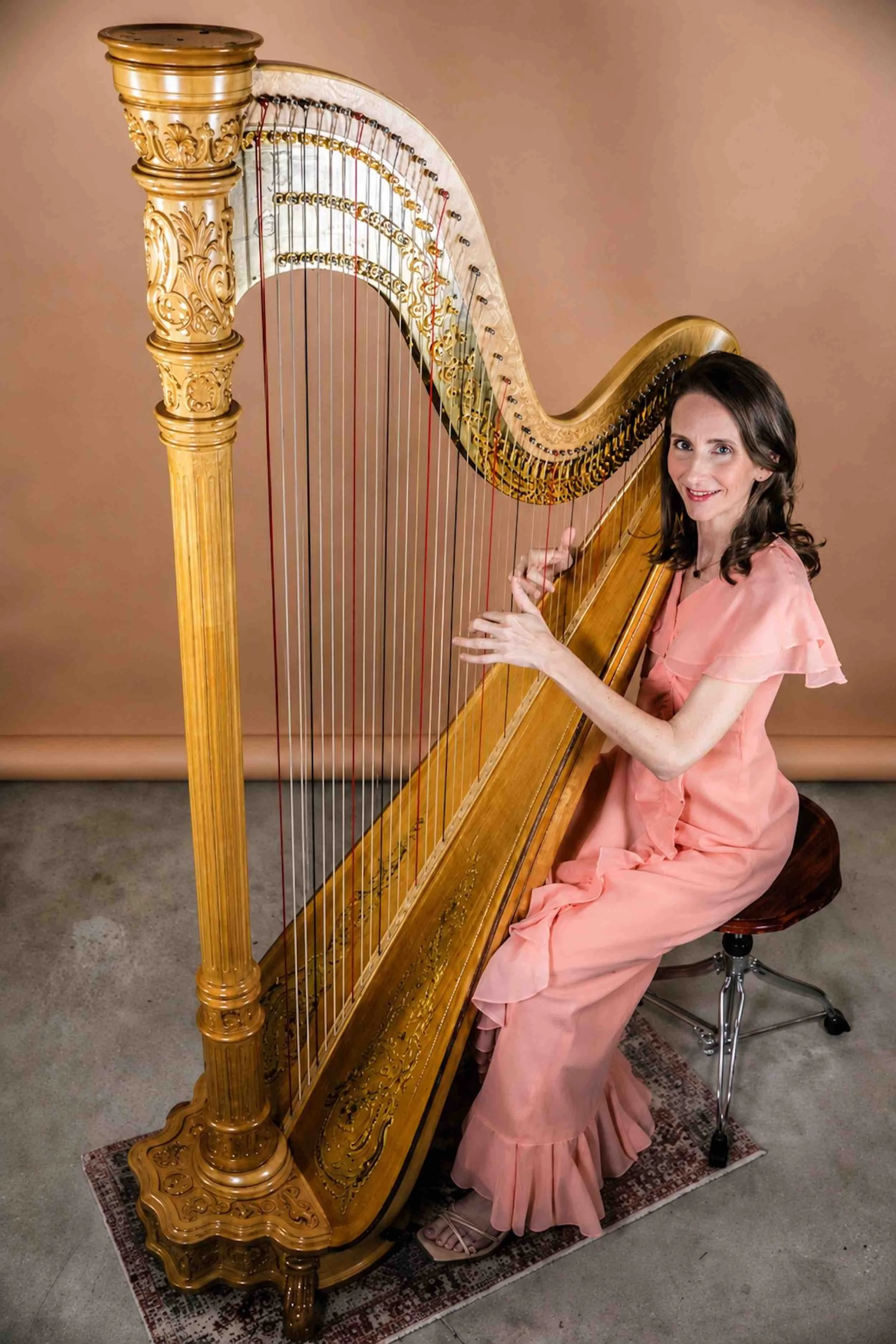 A woman in a soft pink dress sitting on a stool playing a large concert harp with an ornate carved column, positioned on a small rug.
