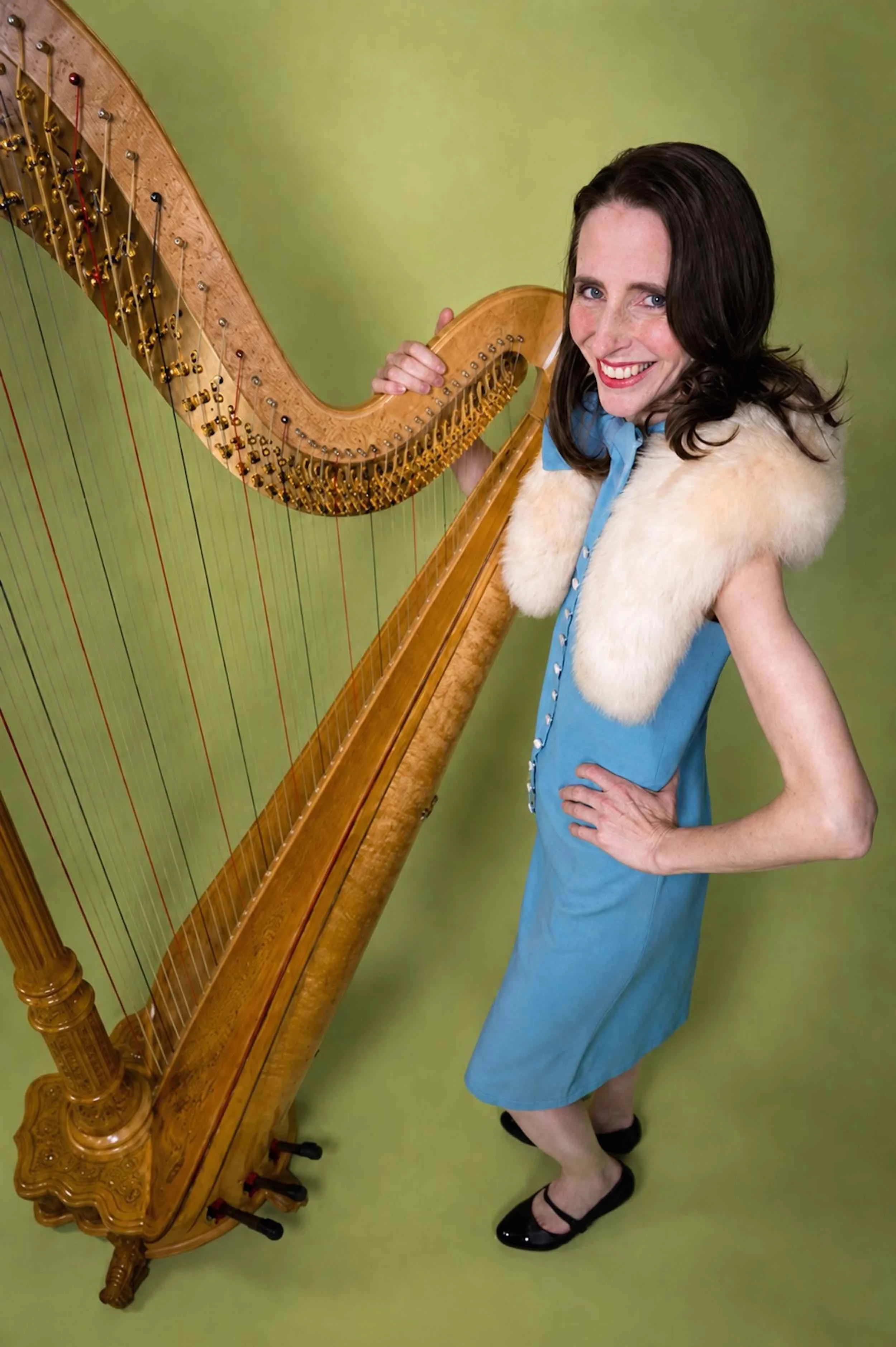 Elegant Woman with dark hair and blue eyes smiling, wearing a blue dress with a faux fur stole, standing next to a large concert harp against a green background.