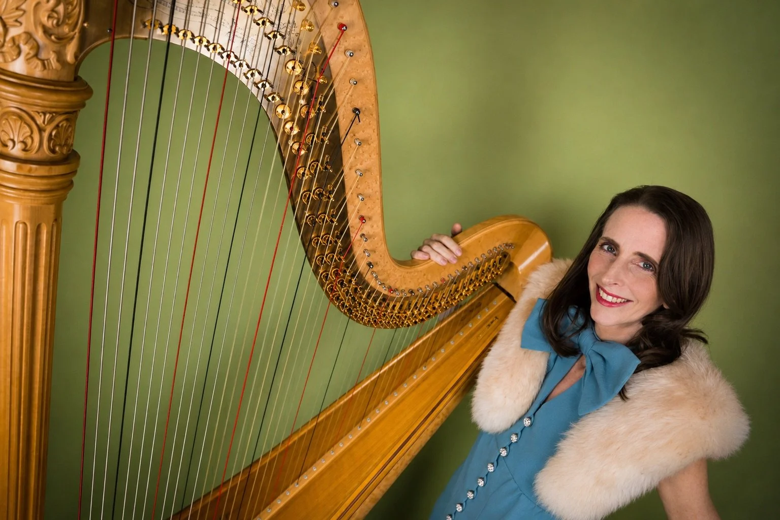 Elegant Woman with dark hair and blue eyes smiling, wearing a blue dress with a faux fur stole, standing next to a large concert harp against a green background.