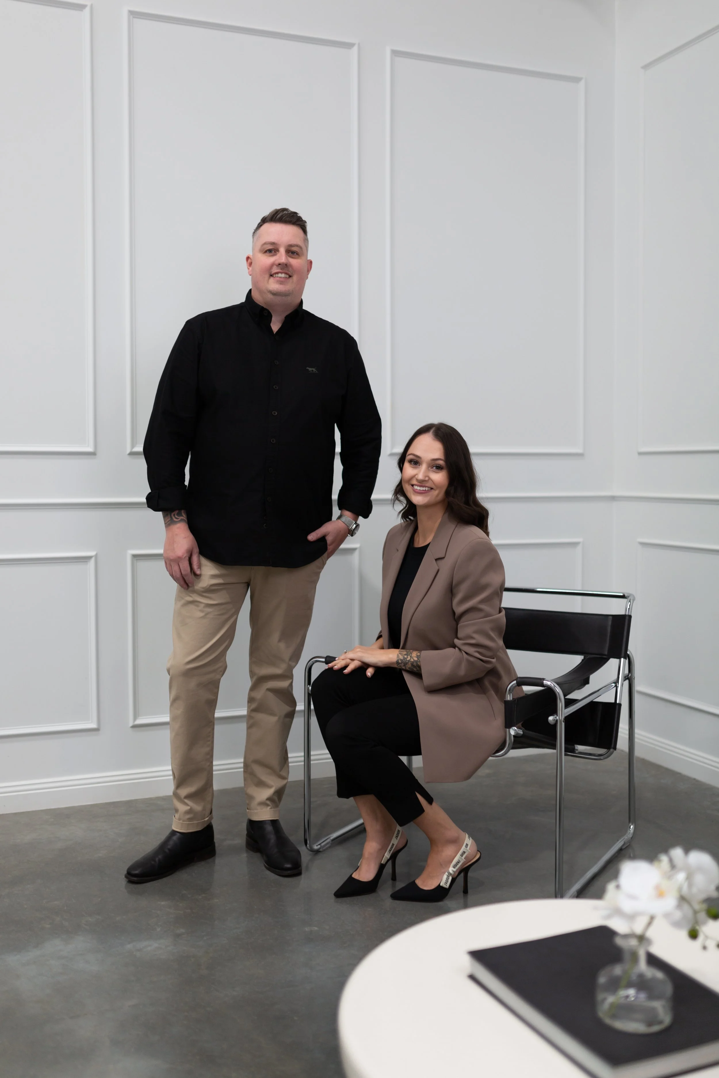 A man in a black shirt and beige pants standing next to a woman seated in a wheelchair, both smiling, in a room with white paneled walls.