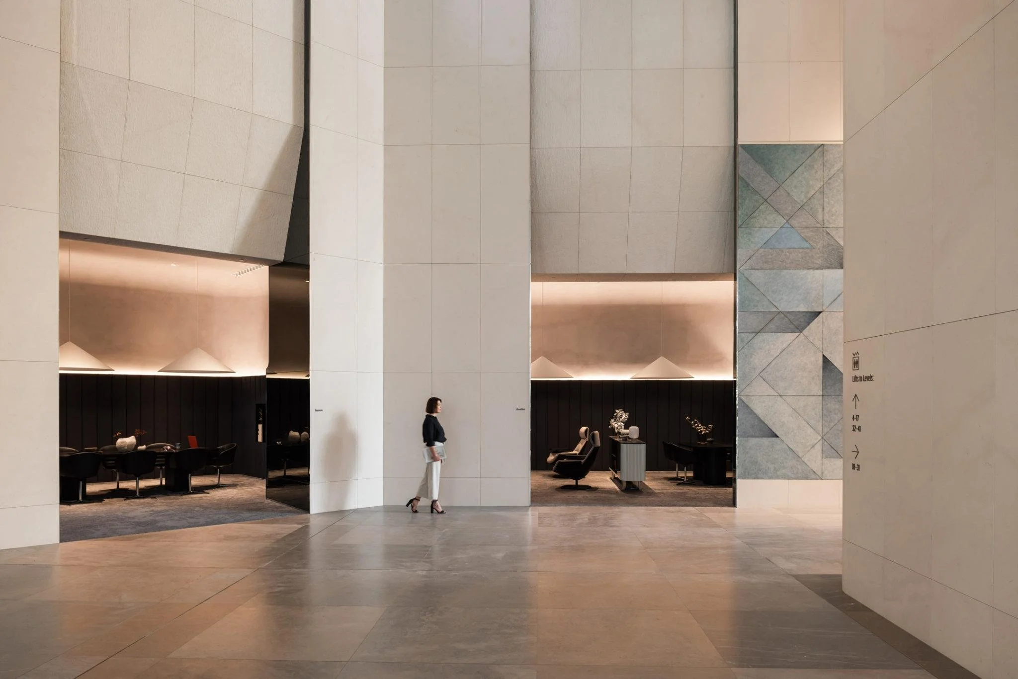 Interior of a modern building with beige tiled walls and floors, and a seating area with black chairs and tables. A woman is walking through the space, carrying a folder or document. There are directional signs on the wall indicating different sections and levels of the building.