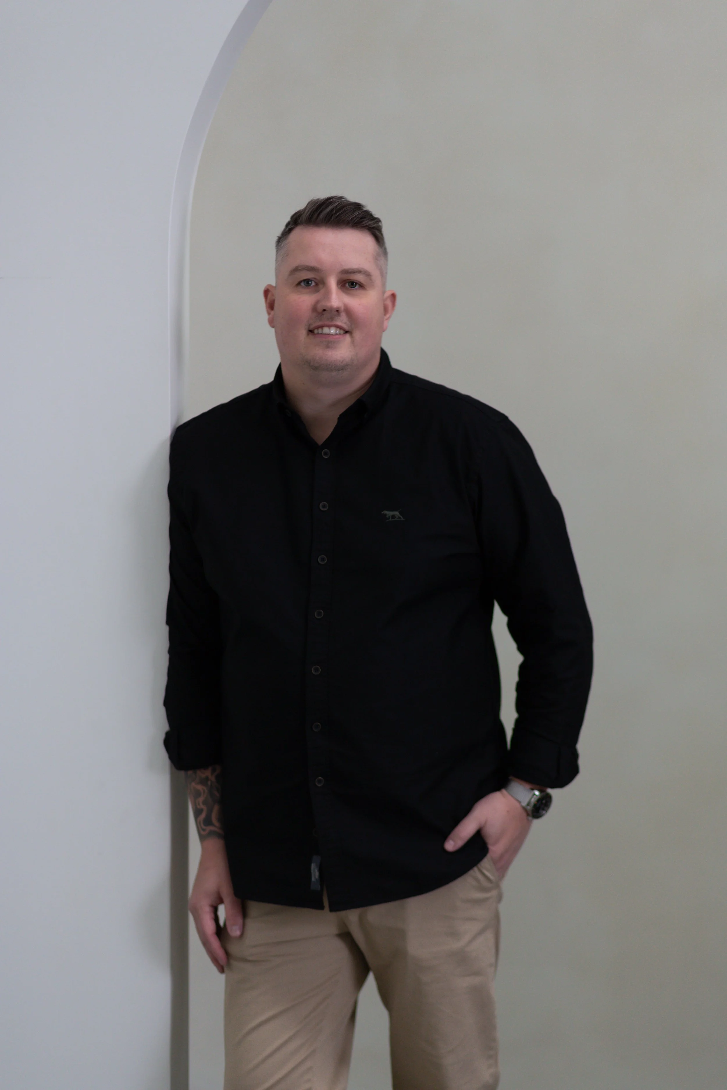 A man standing next to a wall with a geometric design, wearing a black button-up shirt, beige pants, a watch, and his left hand in his pocket.
