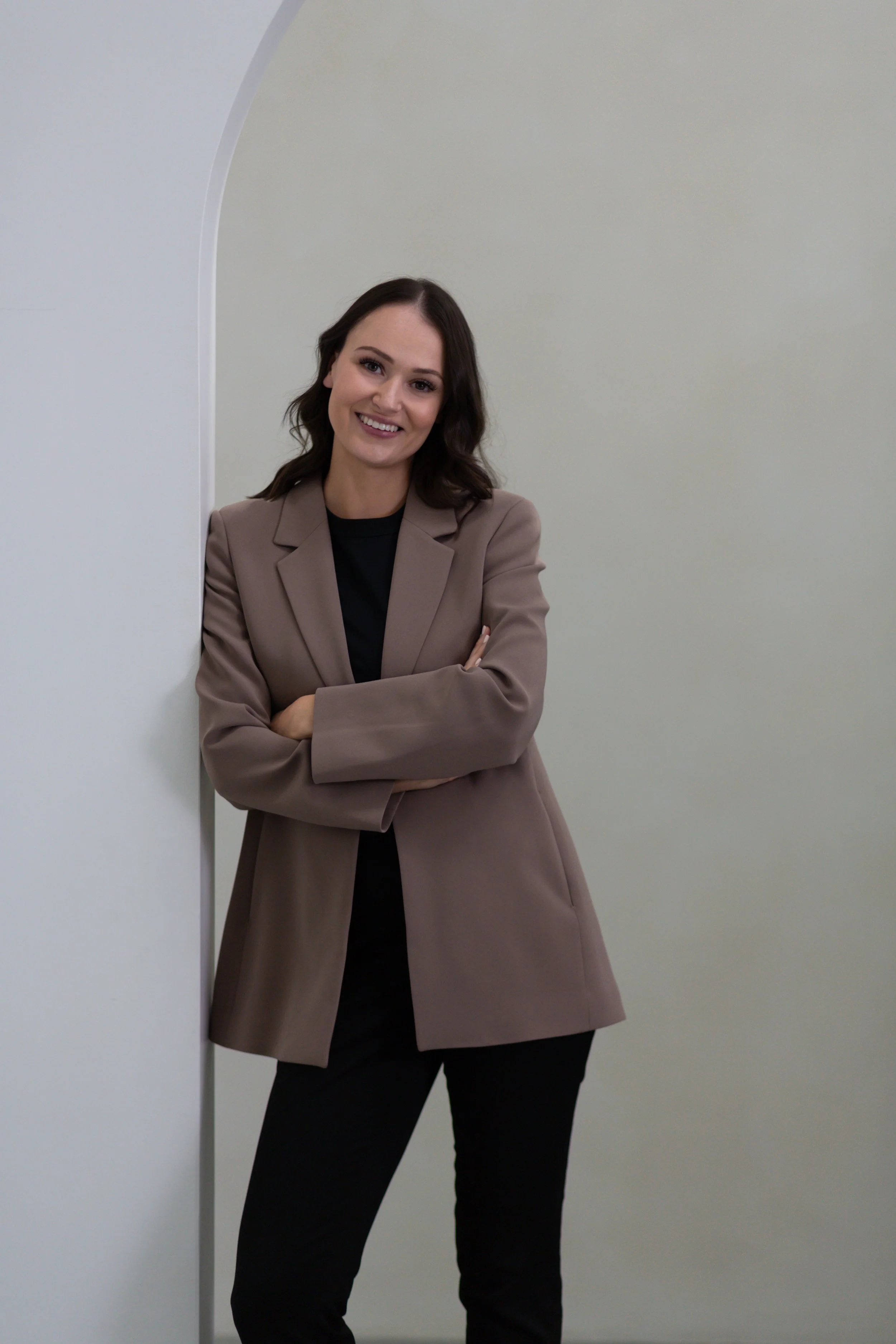 a woman with dark brown hair smiling, wearing a beige blazer and black pants, standing against a plain light-colored wall
