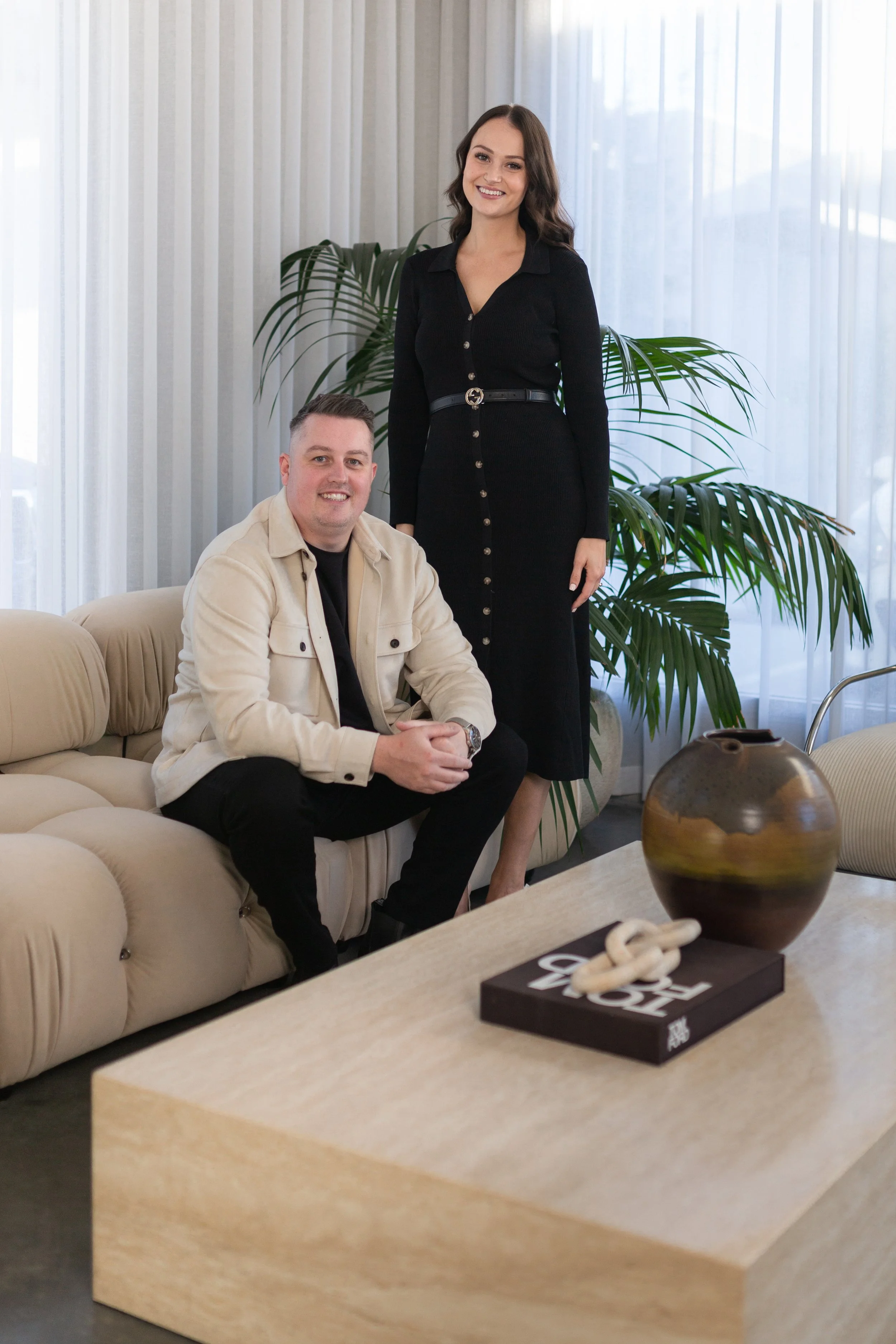 A smiling woman in a black dress standing behind a man seated on a beige sofa. The scene is indoors with light-colored curtains and green plants in the background. There is a decorative pottery piece and a black tray with a white knot-shaped object on a light wooden table in the foreground.