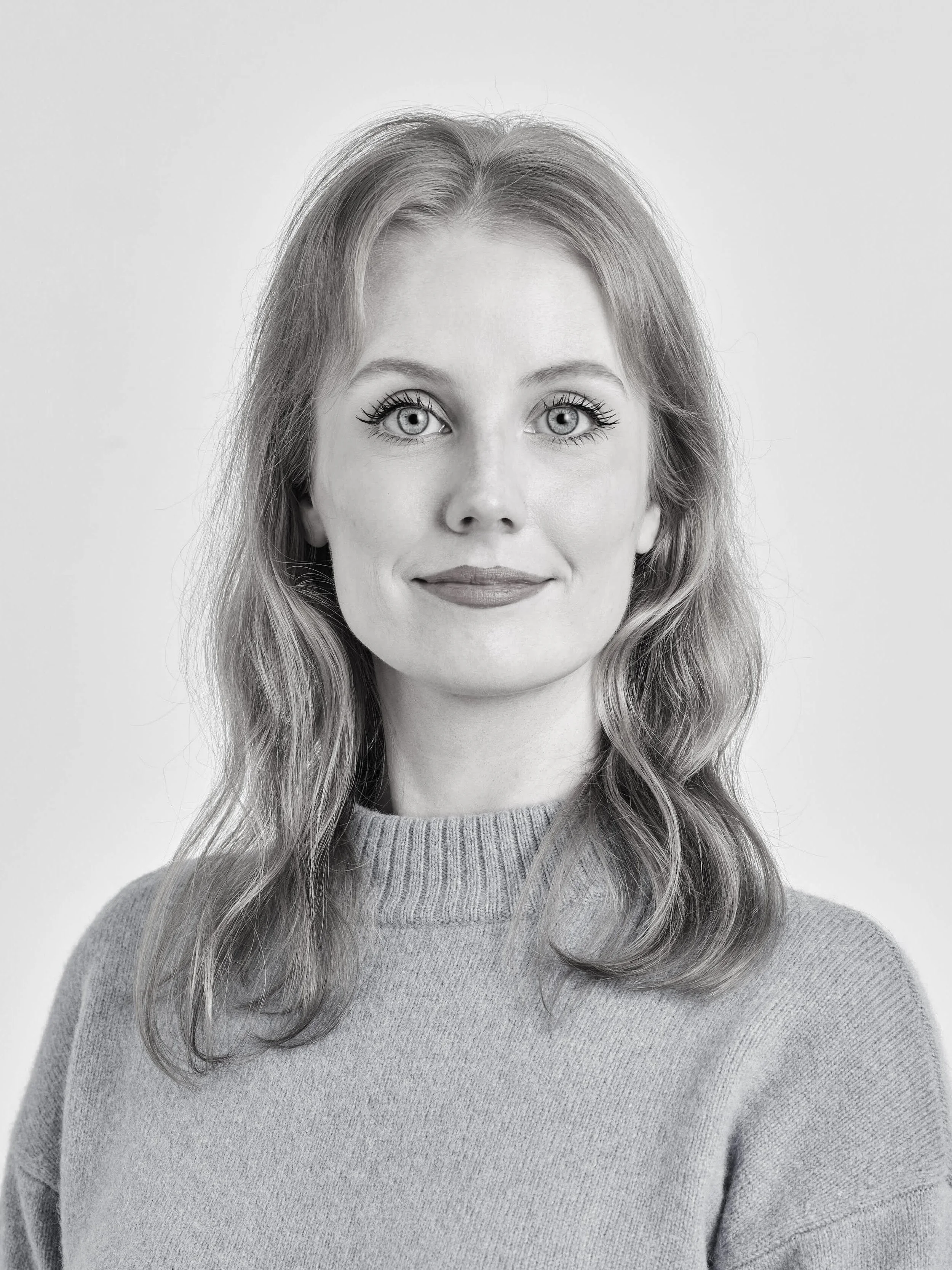 Black and white portrait of a woman with long wavy hair, wearing a sweater, looking directly at the camera with a neutral expression.