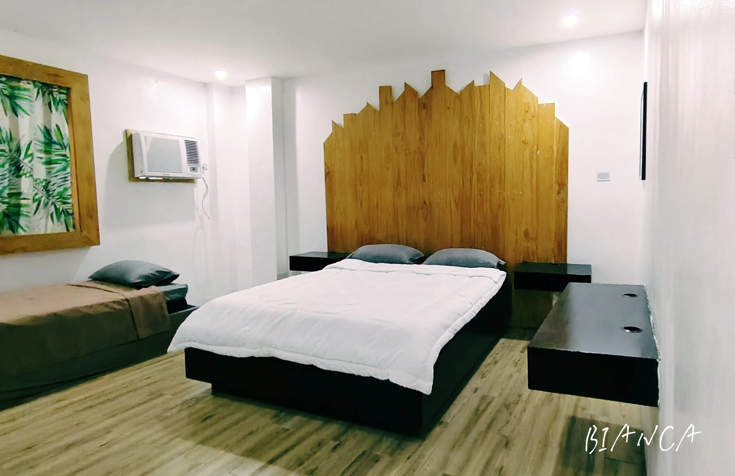 A bedroom with a large bed and pillows, made with a white sheet, and a wooden headboard. There is a smaller mattress with a blanket on it beside the bed. The room has a wooden floor, a window with floral curtains, an air conditioning unit, and a wooden partition.