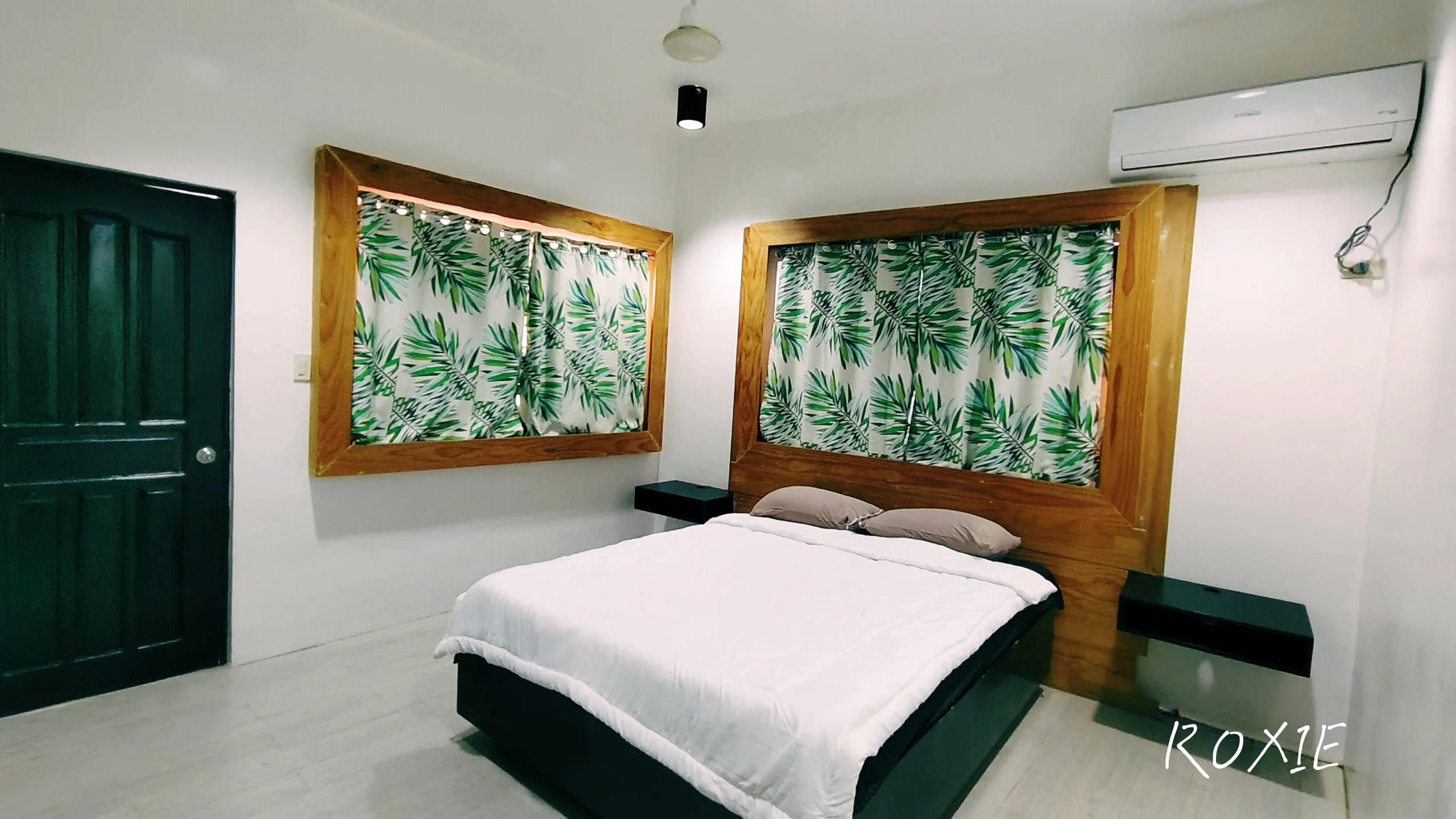 A bedroom with a bed, two side tables, and two windows with leafy patterned curtains, a dark door, a split air conditioning unit, and ceiling lighting.