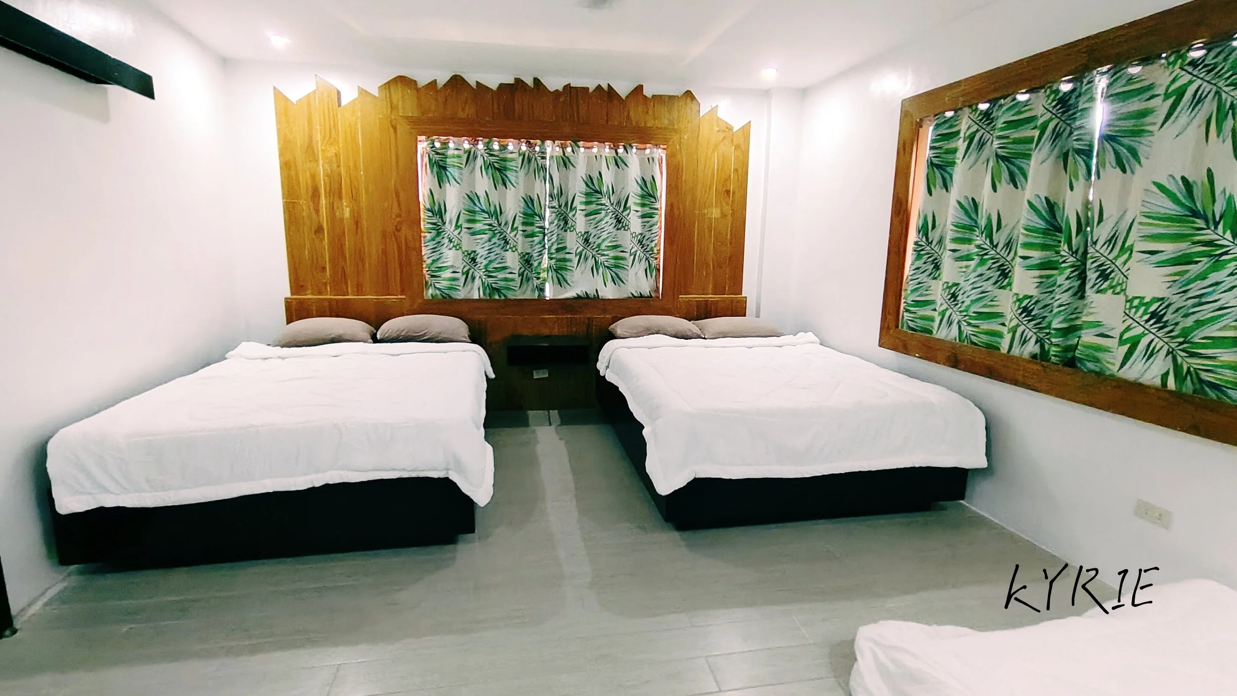 A bedroom with white beds, two pillows each, against a wooden headboard with a leaf pattern behind it, and large leaf-patterned windows on both sides.
