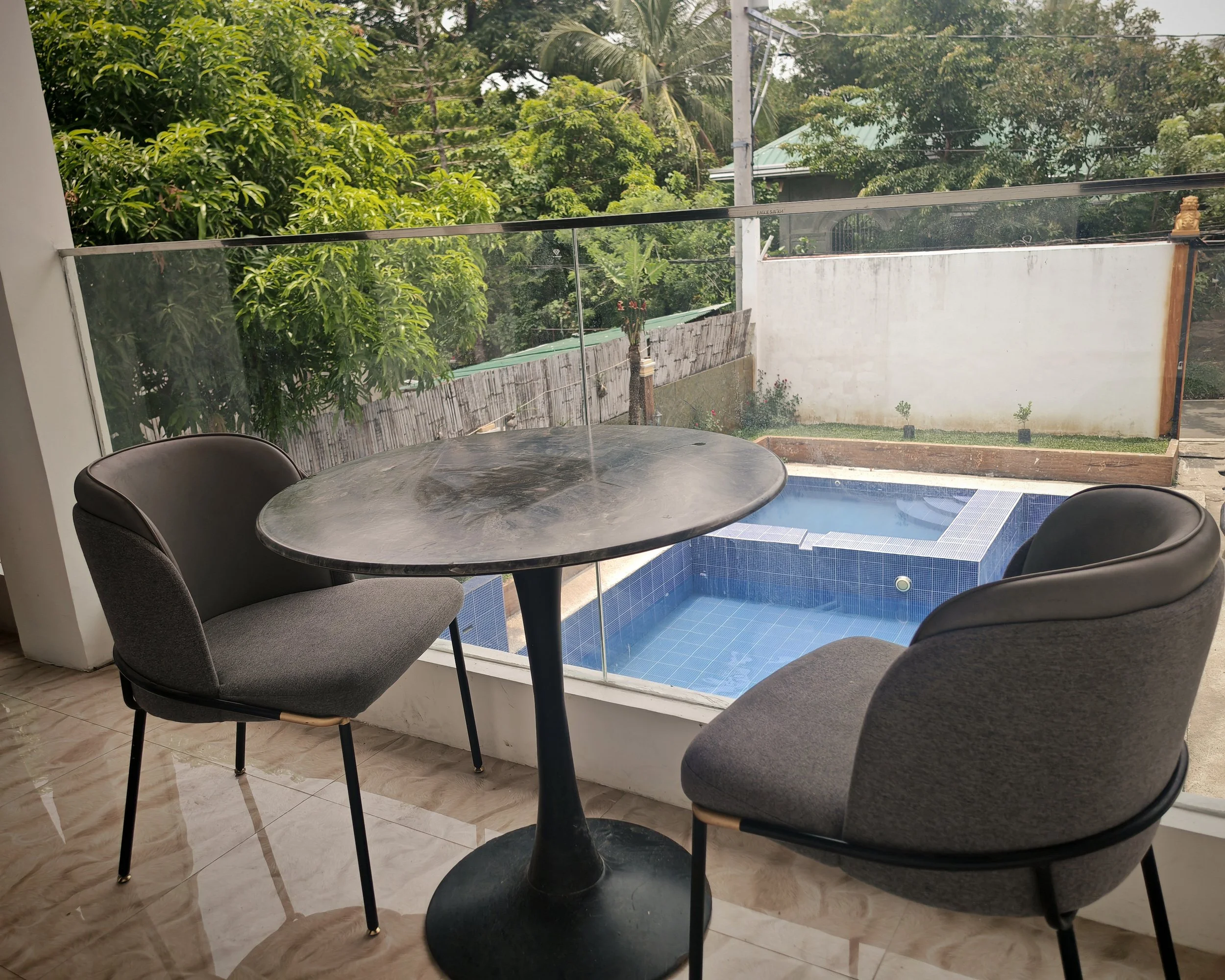 A round black table with two gray chairs situated on a tiled floor inside a room. The room has large glass windows with a view of a backyard that contains a small pool, a white wall, a bamboo fence, and lush green trees and plants outside.