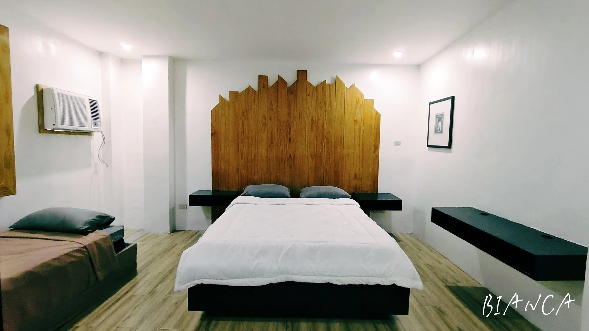 A bedroom with a white bed, gray pillows, a wooden headboard, and black side tables. There is an air conditioning unit on the wall, framed picture on the right wall, and another bed with a brown blanket on the left.