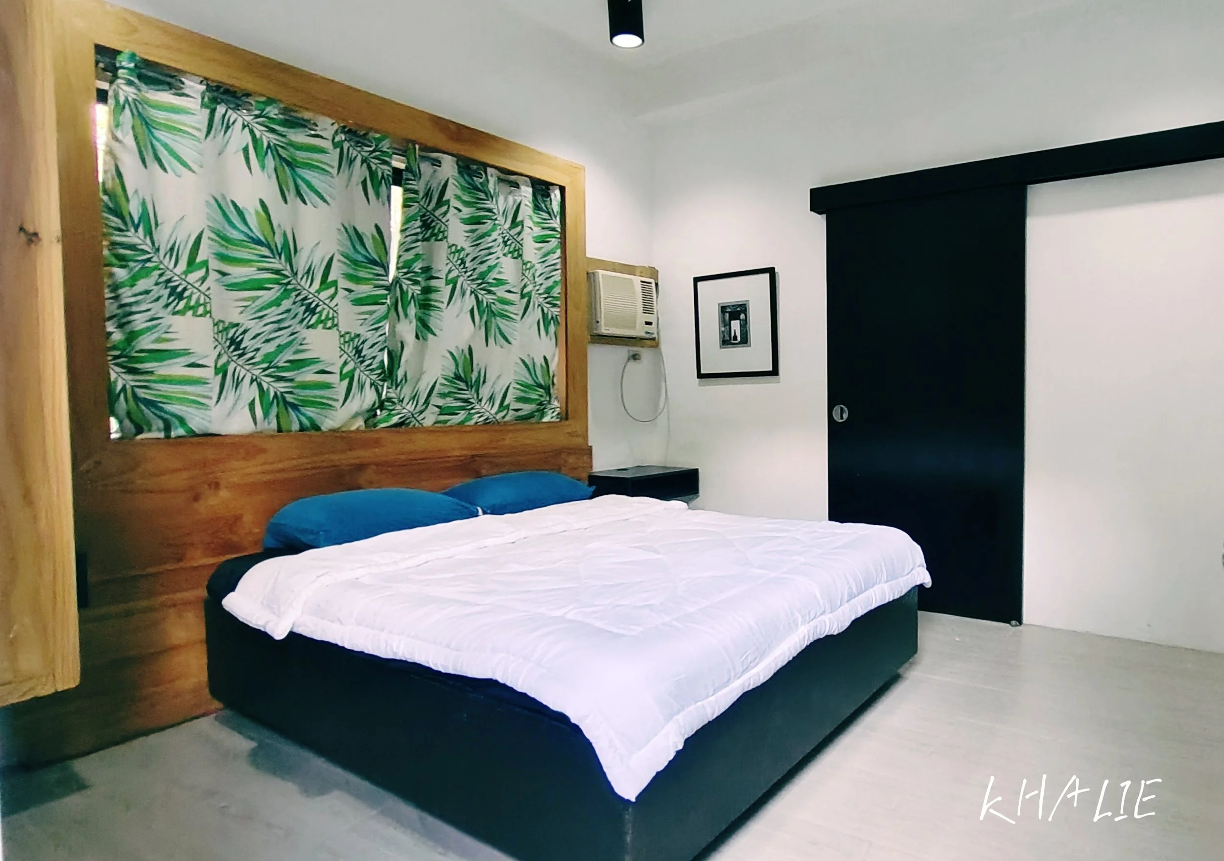 A bedroom with a wooden headboard, a white bed with black and blue bedding, tropical leaf-patterned curtains, an air conditioning unit on the wall, framed artwork, and a black sliding door.