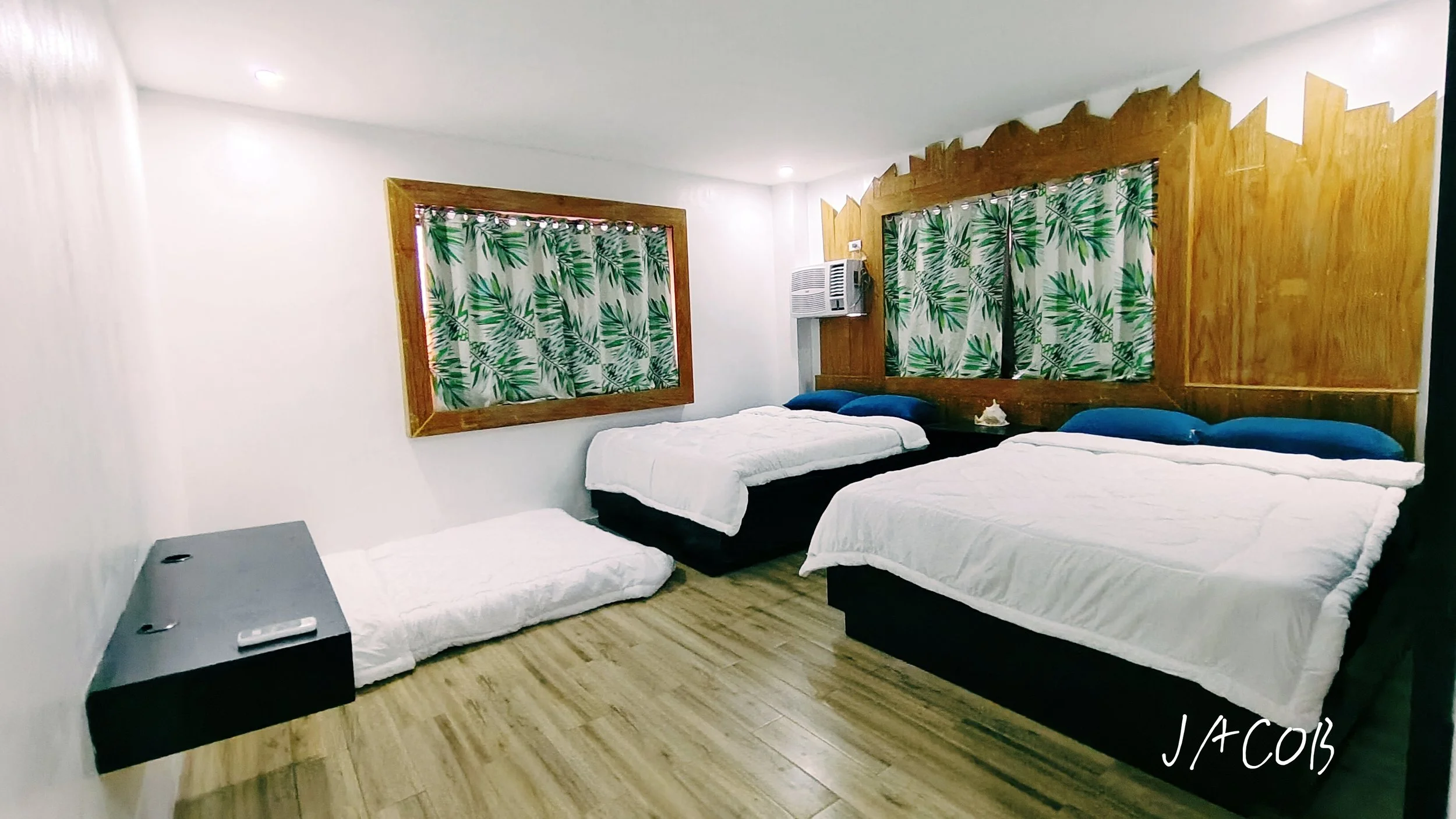 A hotel room with three beds, two with blue pillows and one without a pillow, white bed coverings; a window with tropical leaf-patterned curtains, wooden headboards and trim, a small black nightstand, and an air conditioning unit on the wall.