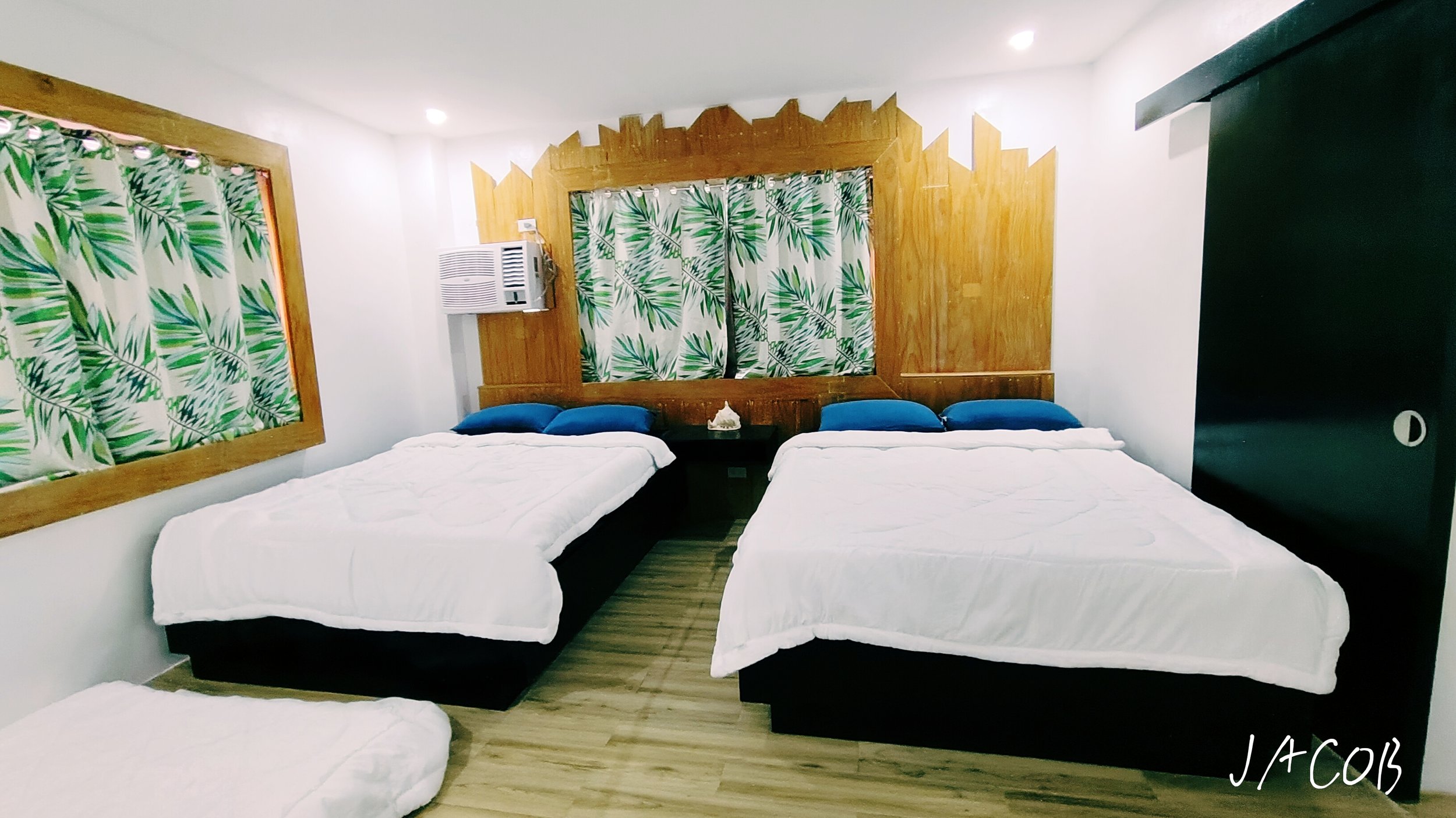 A hotel room with three beds, each with white bedding and blue pillows. The wall behind the beds has wooden paneling and tropical leaf-patterned curtains. An air conditioning unit is mounted on the wall near the center bed, and the floor is wood.