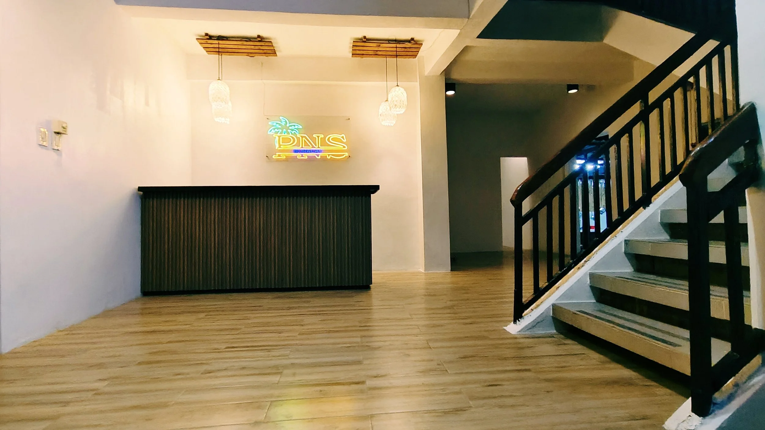 Interior view of a modern space with wooden flooring, a staircase with black railing, a black bar or counter with corrugated metal front, illuminated pendant lights, and a neon sign with a palm tree and the words 'DLS' and 'Surf' on the wall.