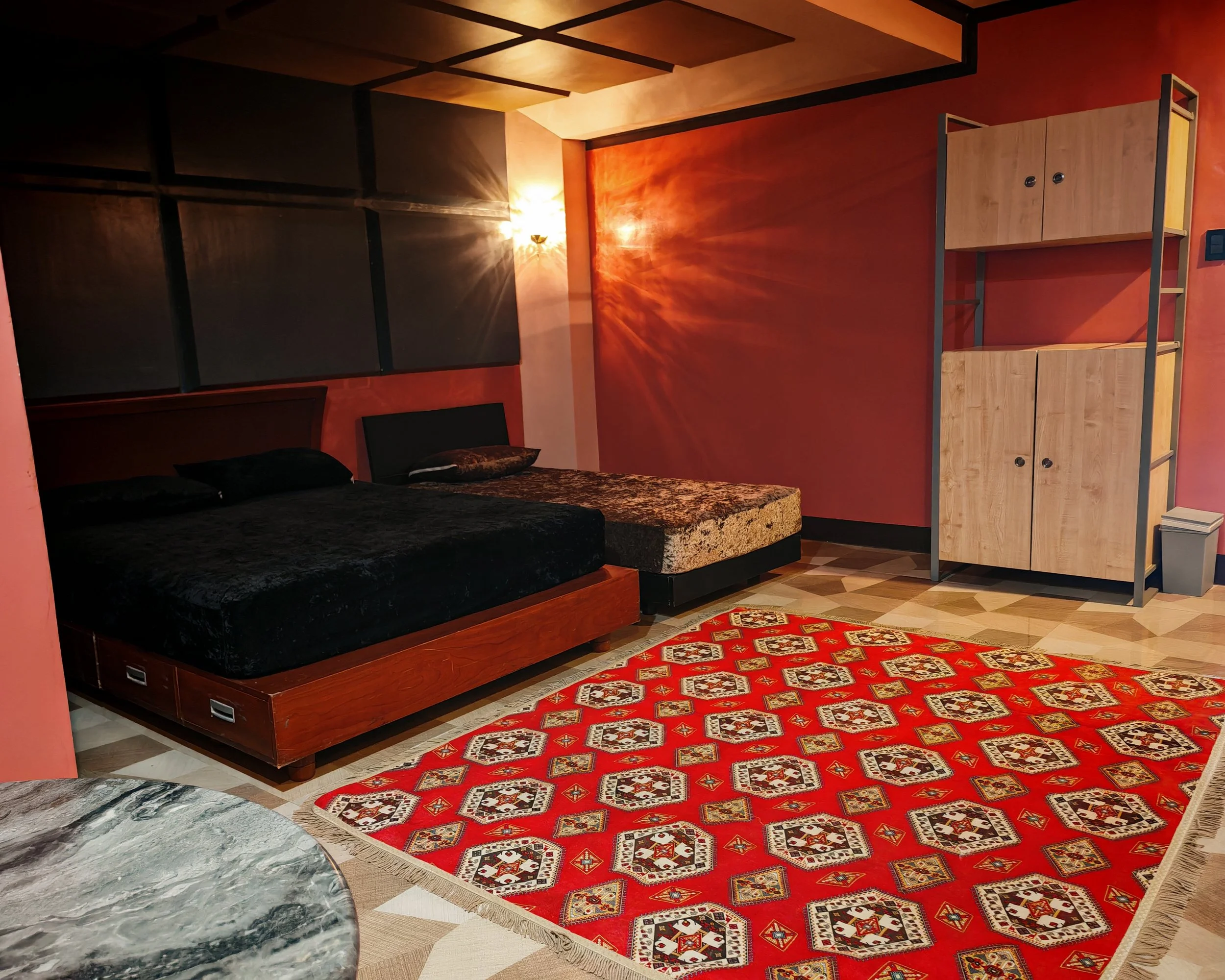 A bedroom with a maroon wall, a bed with black bedding, a red patterned rug, a wooden shelf with two doors, and a marble surface in the foreground