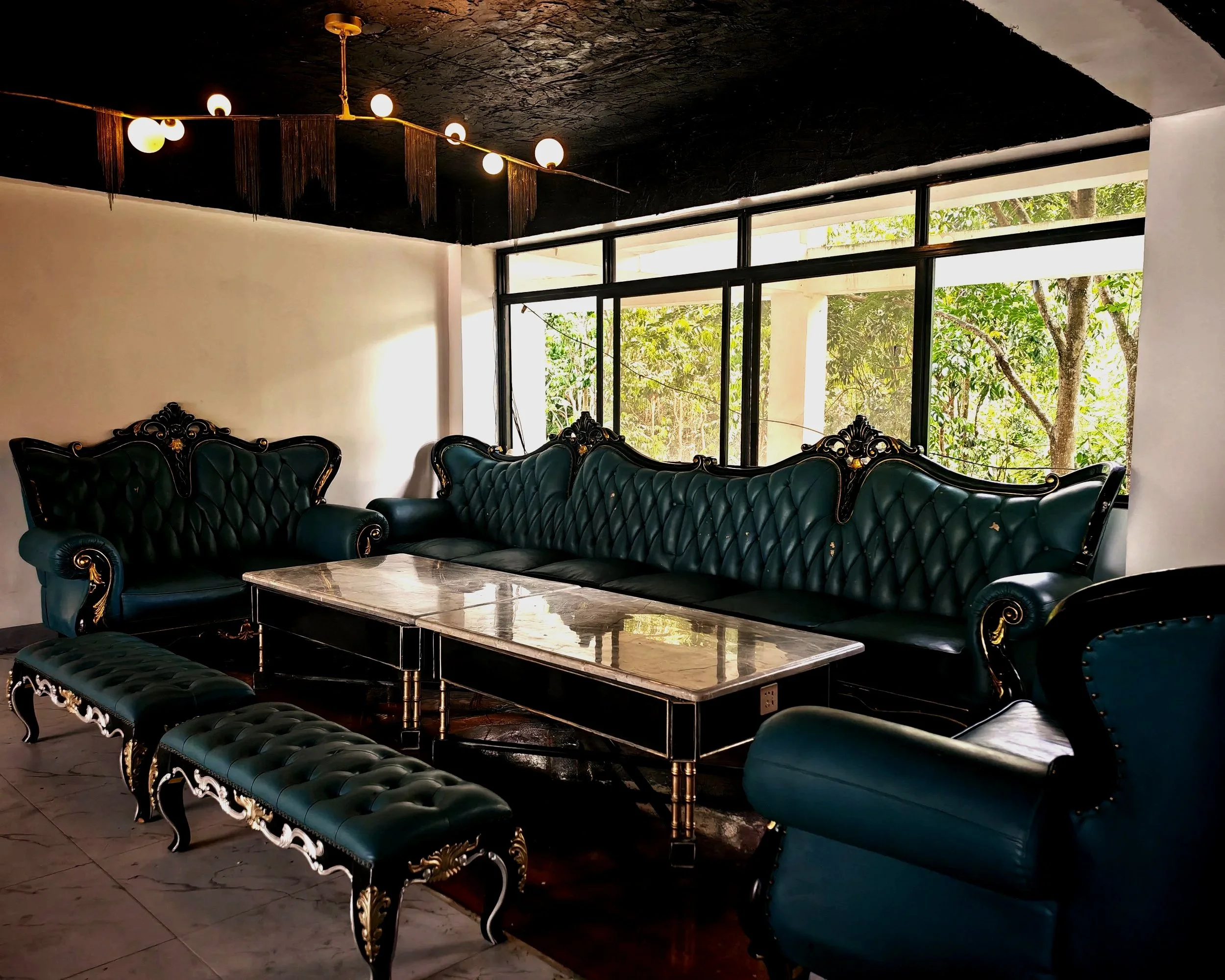 Living room with black tufted leather sofas with ornate gold and wood accents, a marble coffee table, large window with a view of trees outside, and a ceiling with hanging string lights.